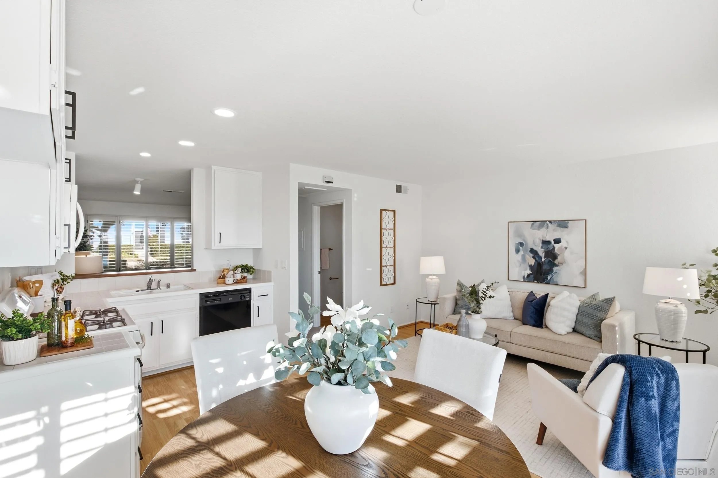 Open-plan kitchen and living room with a wood dining table, white cabinetry, and sunlight across the floor.