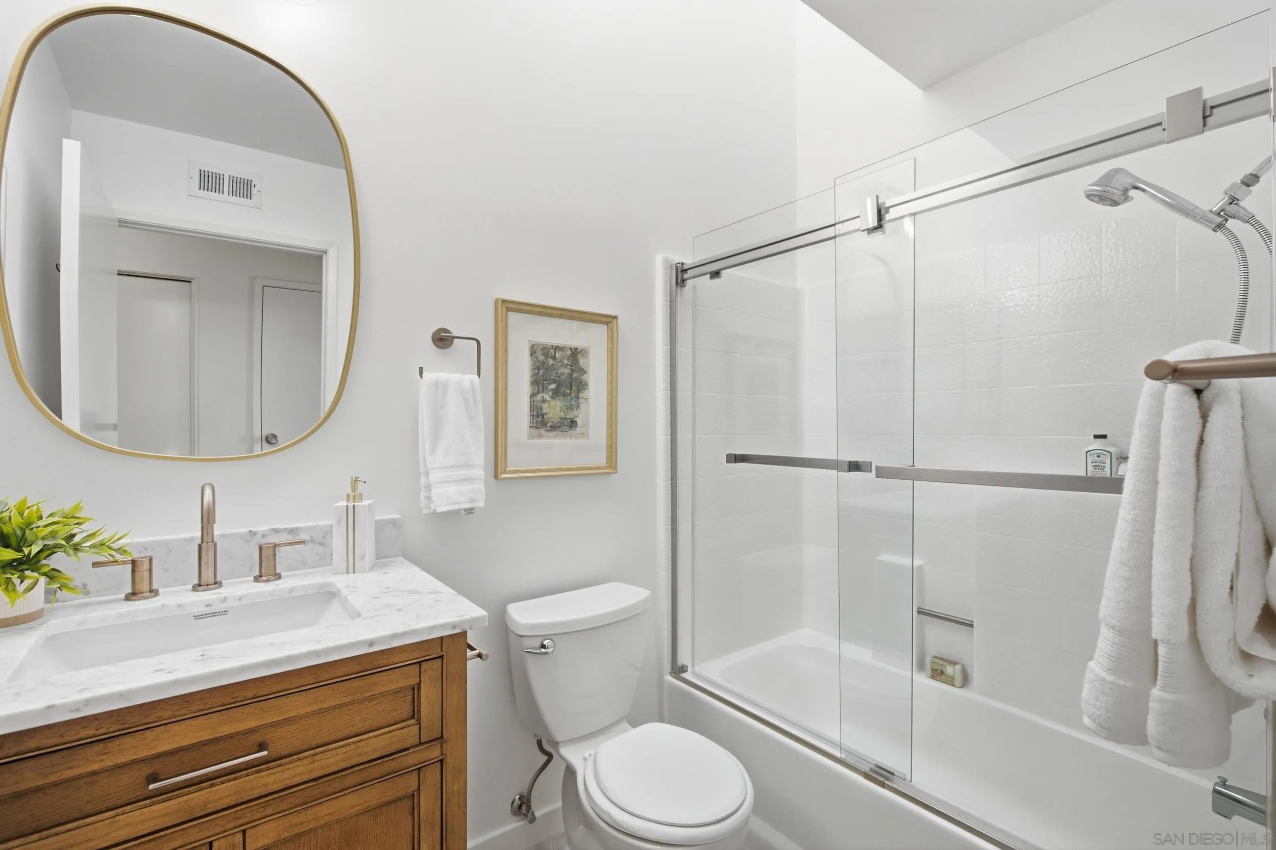 Bright bathroom with a wood vanity, gold-framed mirror, and glass-enclosed tub and shower.