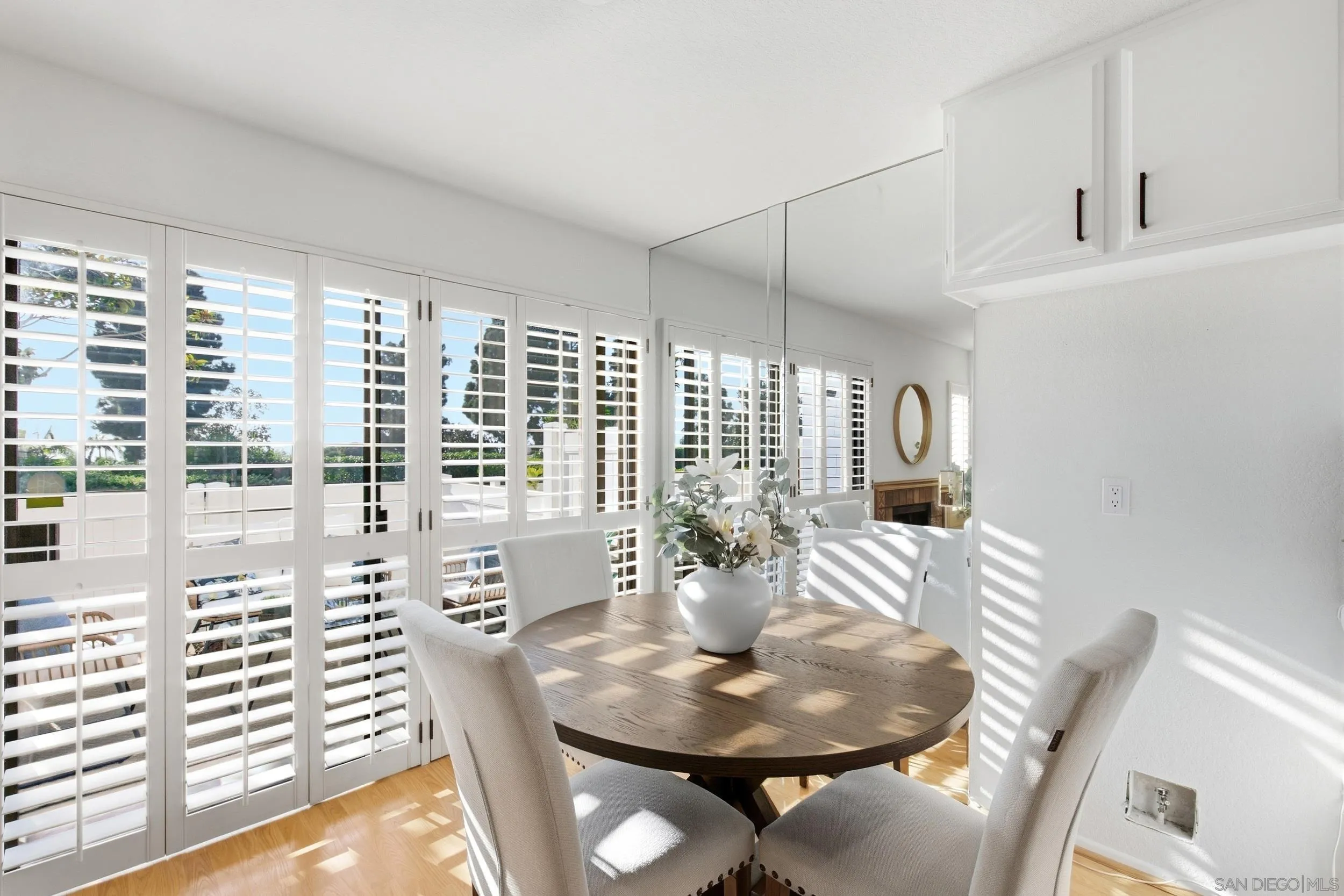 Staged dining room with a round wood table and sunlight filtering through white plantation shutters.