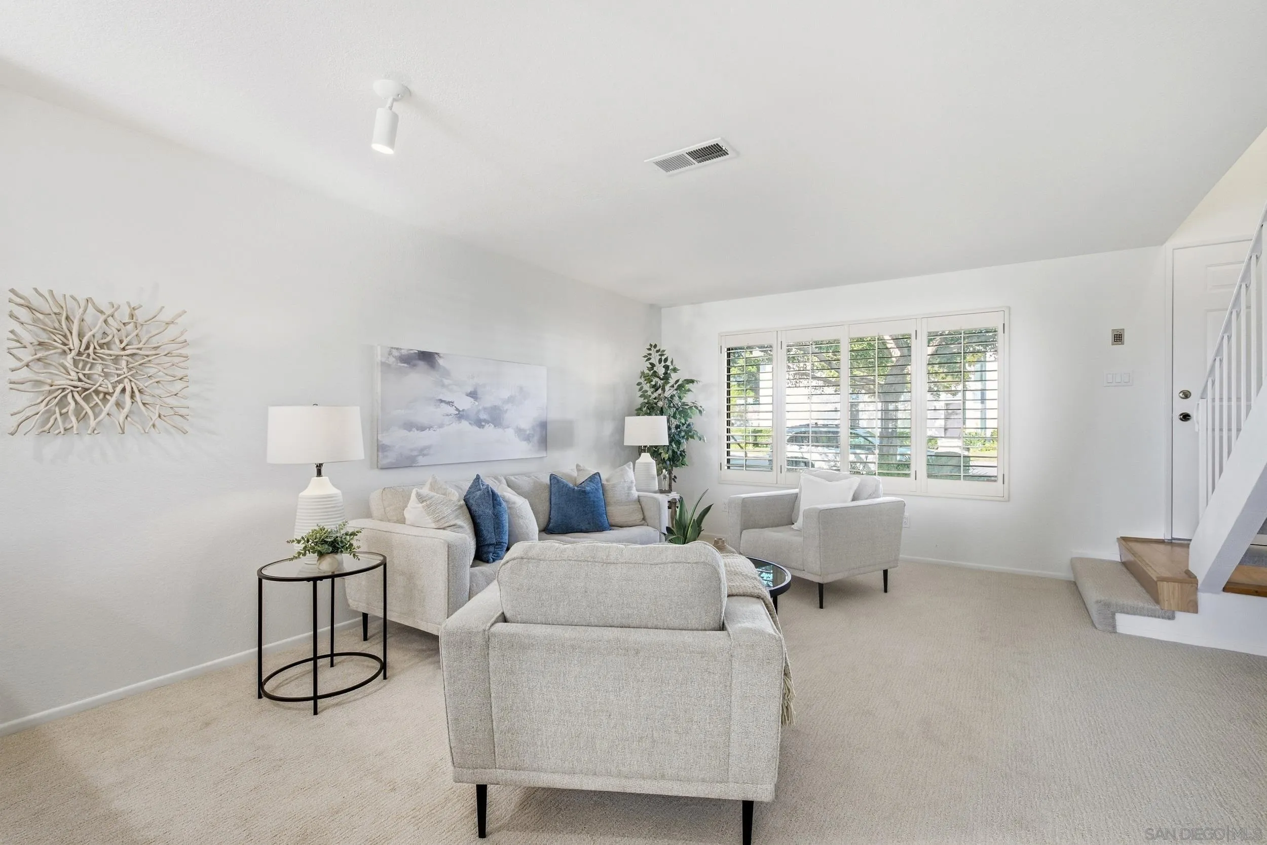 Staged living room with neutral seating, blue accent pillows, shuttered windows, and a staircase.