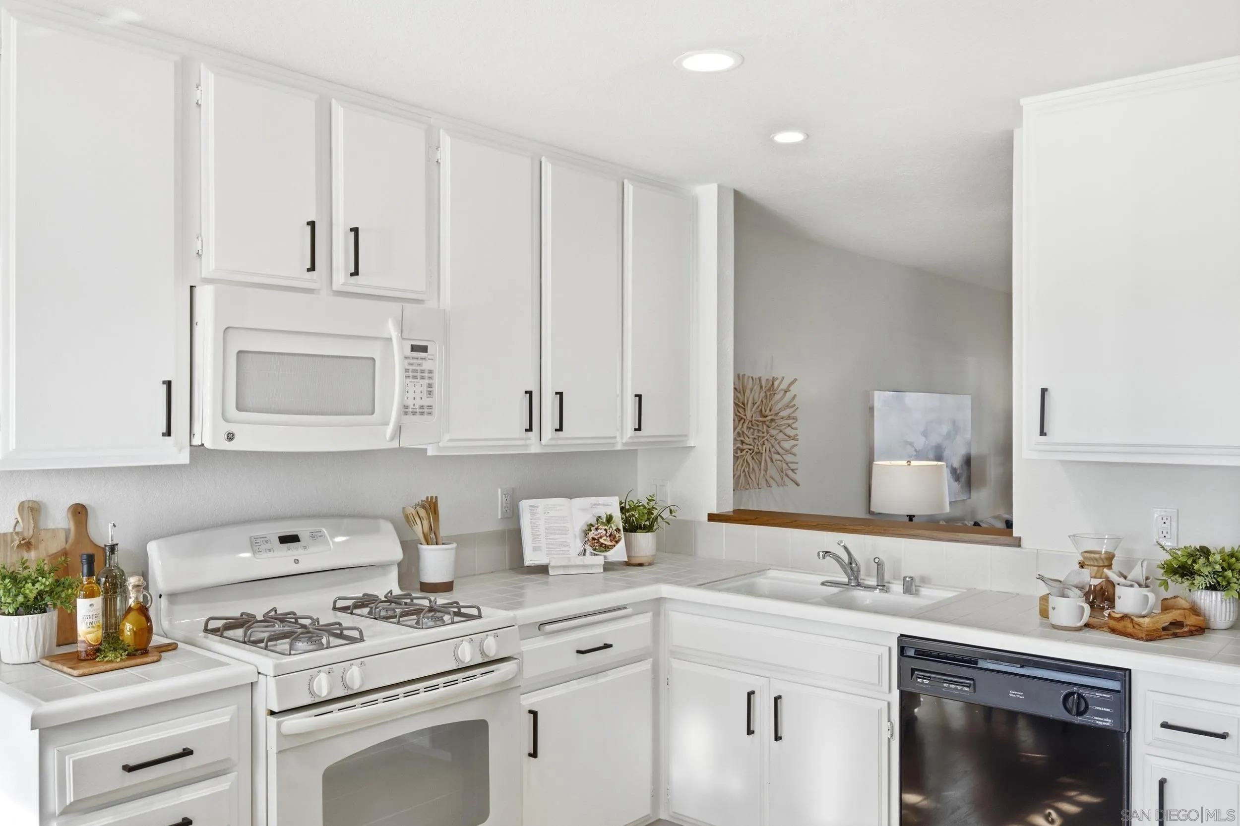 Bright white kitchen with a gas range, gray tile counters, and a pass-through to the adjoining room.