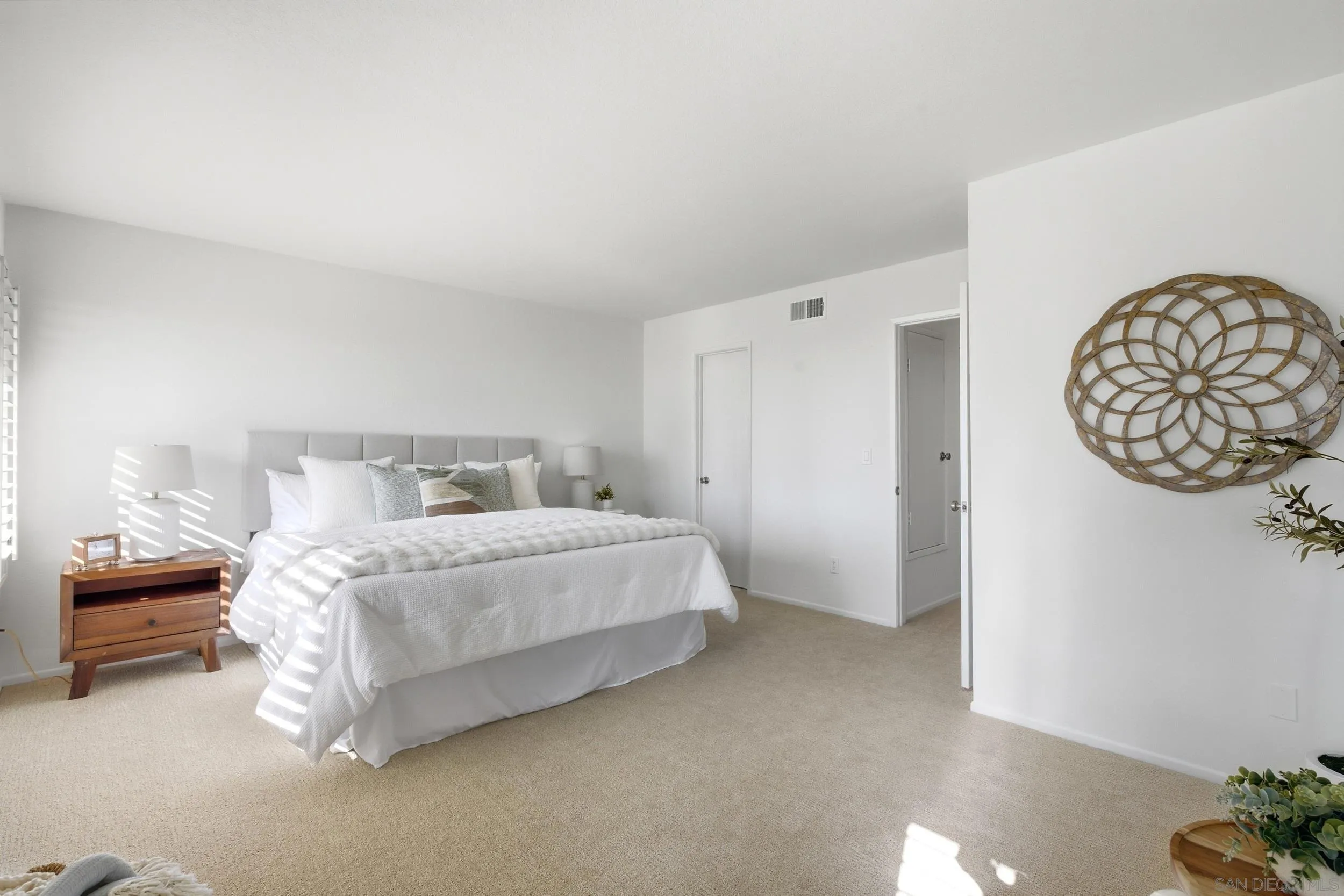 Bright staged bedroom with a white bed, beige carpet, and sunlight streaming through blinds.