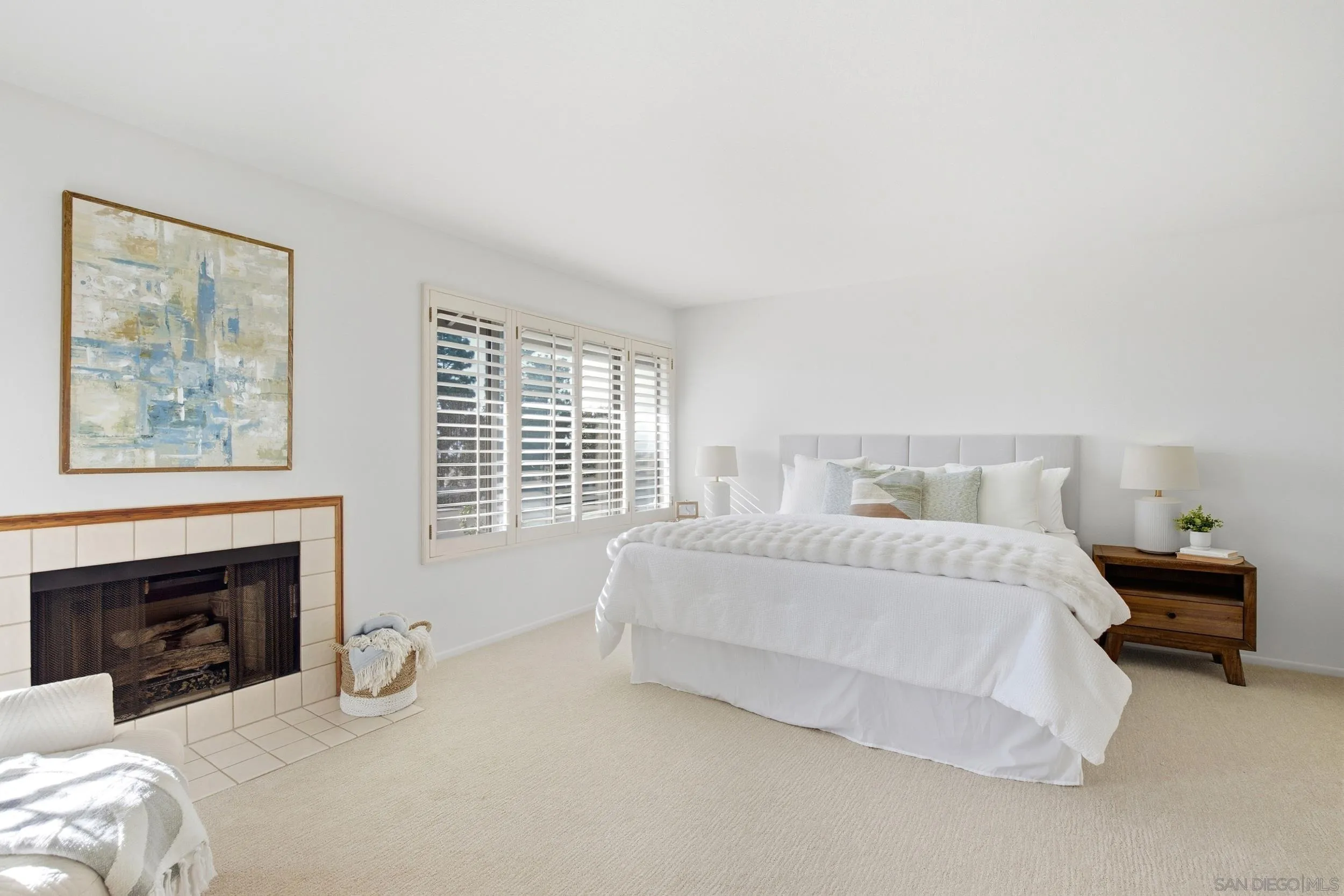 Staged bedroom with a white bed, plantation shutters, and a tiled fireplace.