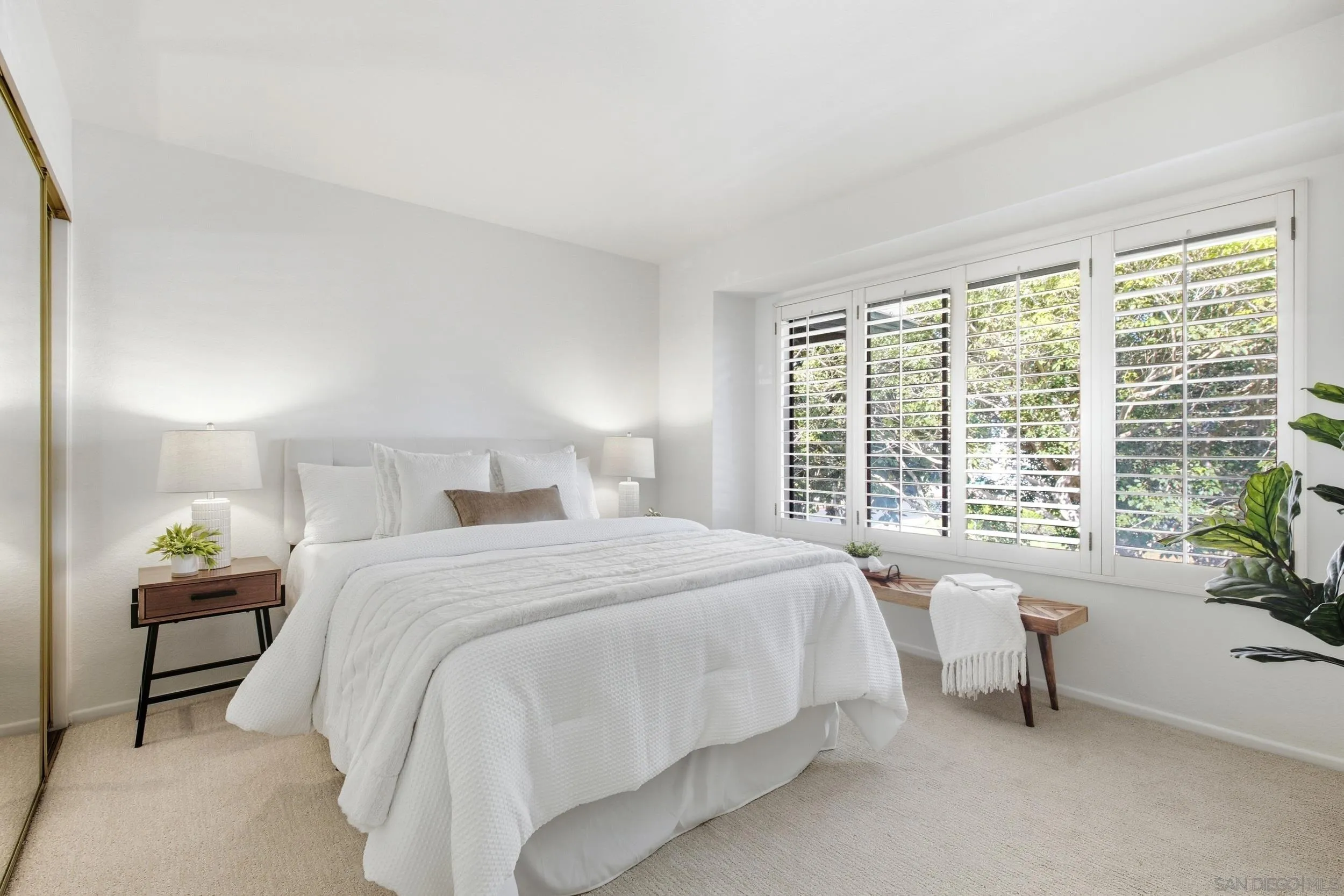 Staged bedroom with a white bed and wide plantation-shuttered windows overlooking leafy trees.