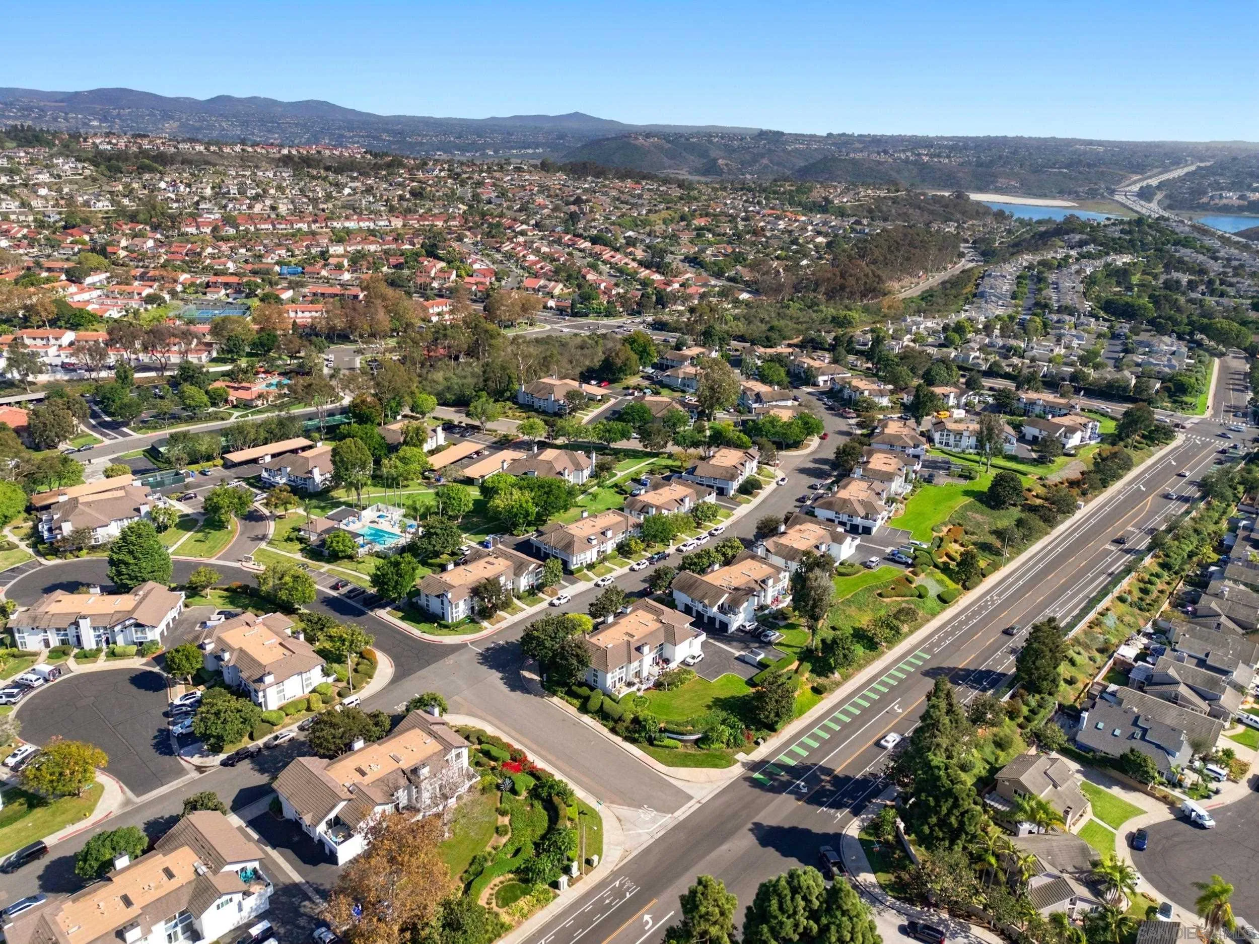 Aerial view of a residential neighborhood with a pool, major road, hills, and reservoir in the distance.