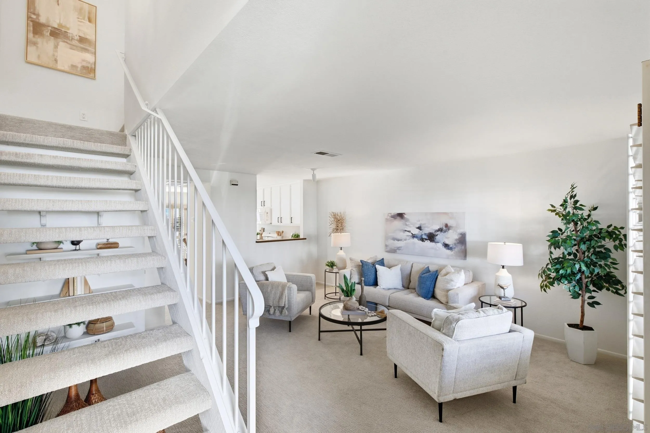 Staged living room with a carpeted staircase, neutral seating, and blue accent pillows.