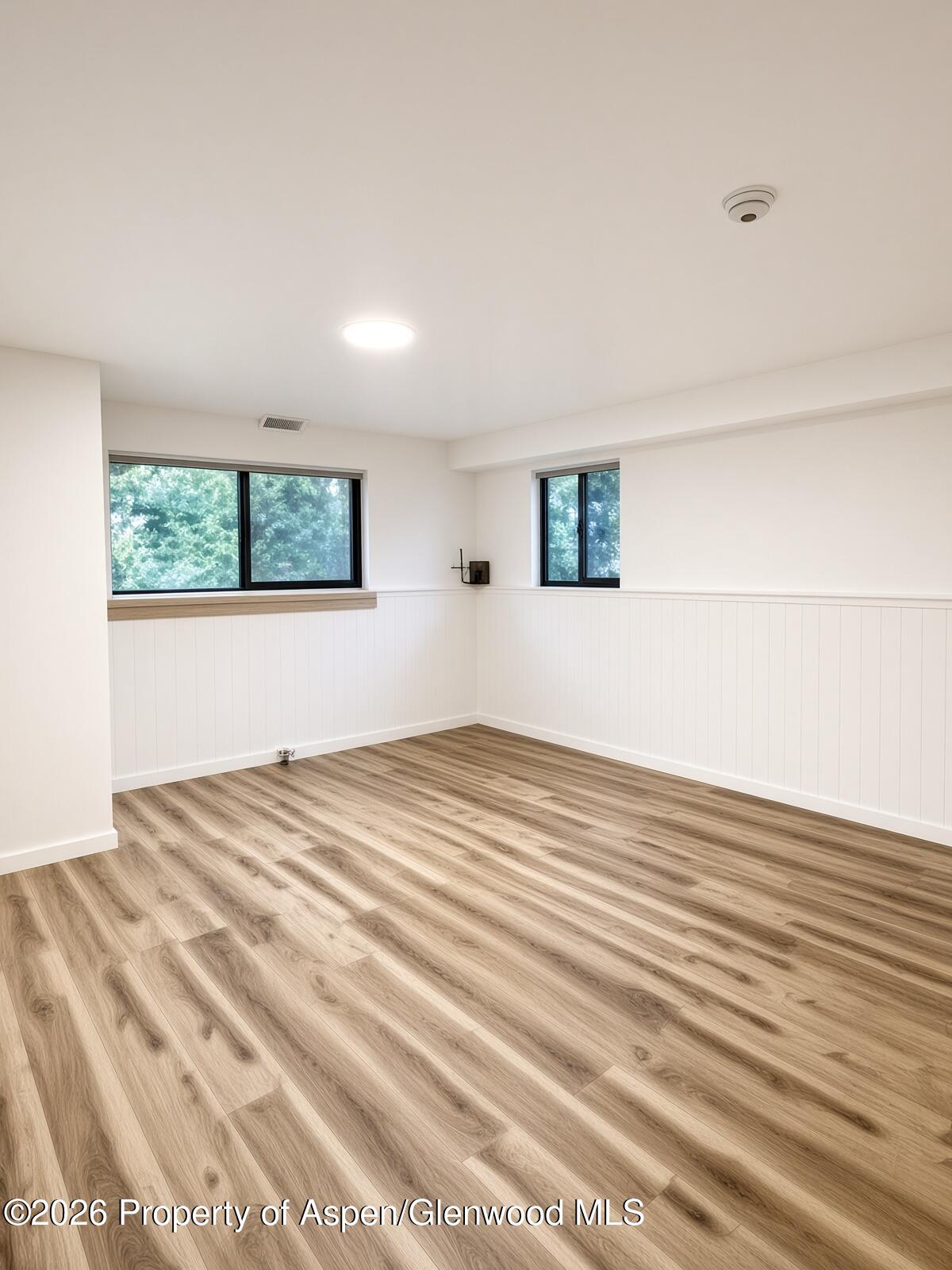 Empty room with white paneled walls, two windows, and light wood-look plank flooring.