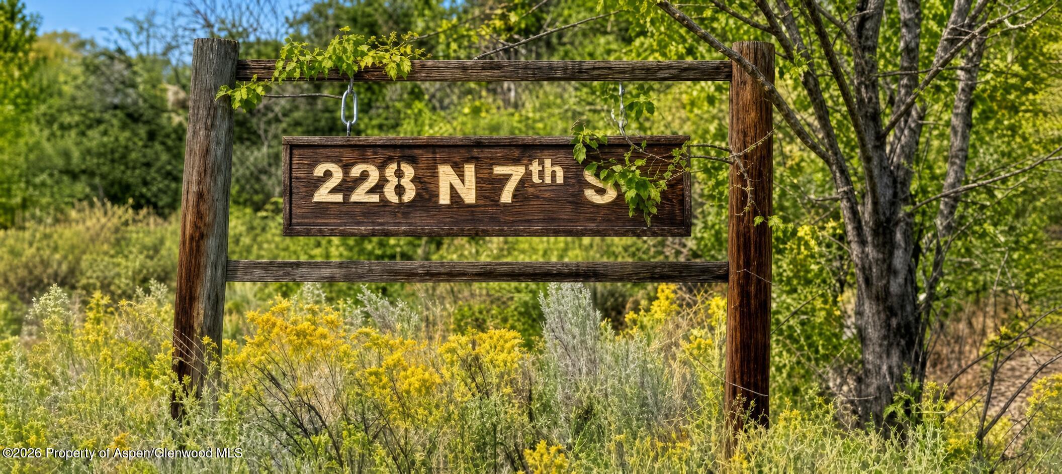 228 N 7th Street address sign hanging from a wooden frame among trees and wildflowers.