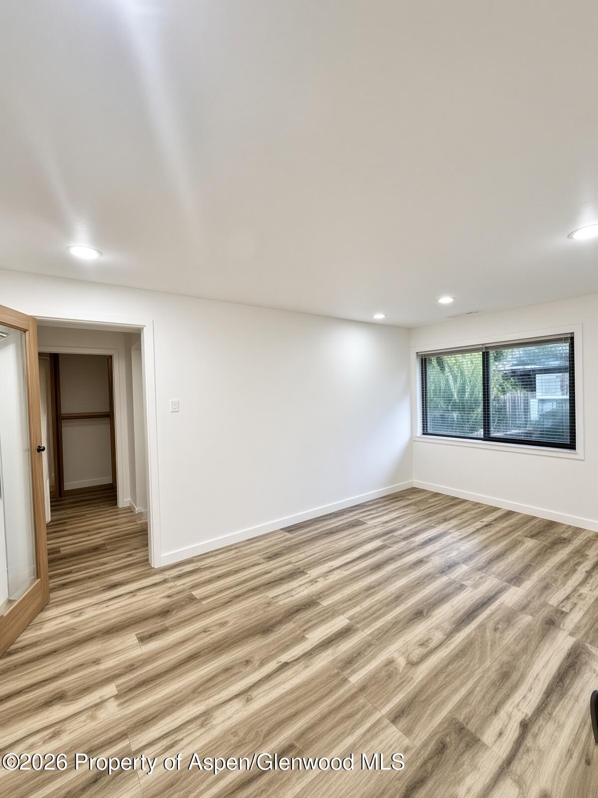 Empty bedroom with wood-look plank flooring, recessed lights, and a wide window with blinds.