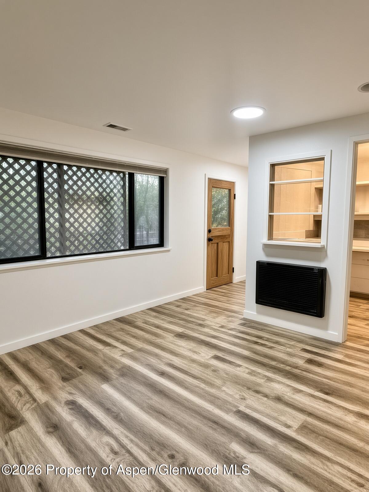 Unfurnished room with plank flooring, a lattice-screened window, wooden door, and wall-mounted heater.