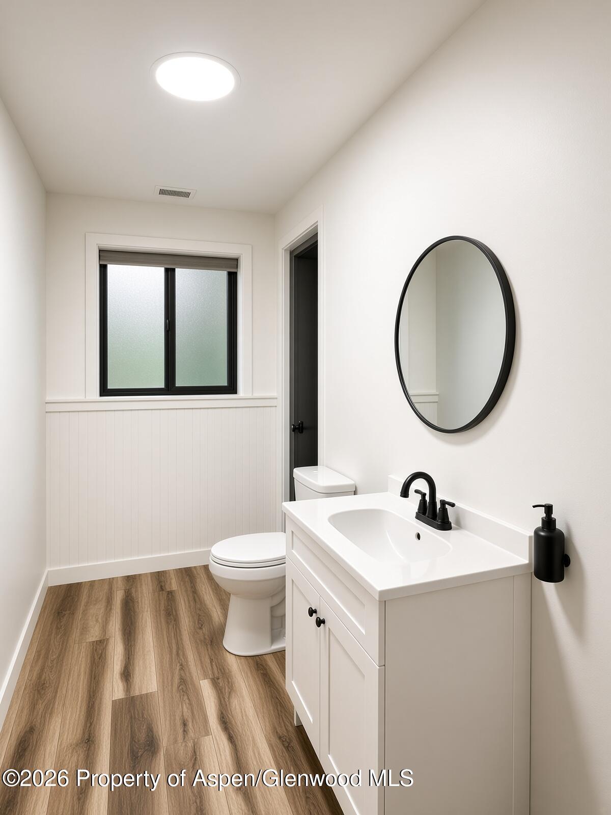 Bathroom with a white vanity, black fixtures, oval mirror, frosted window, and wood-look floor.