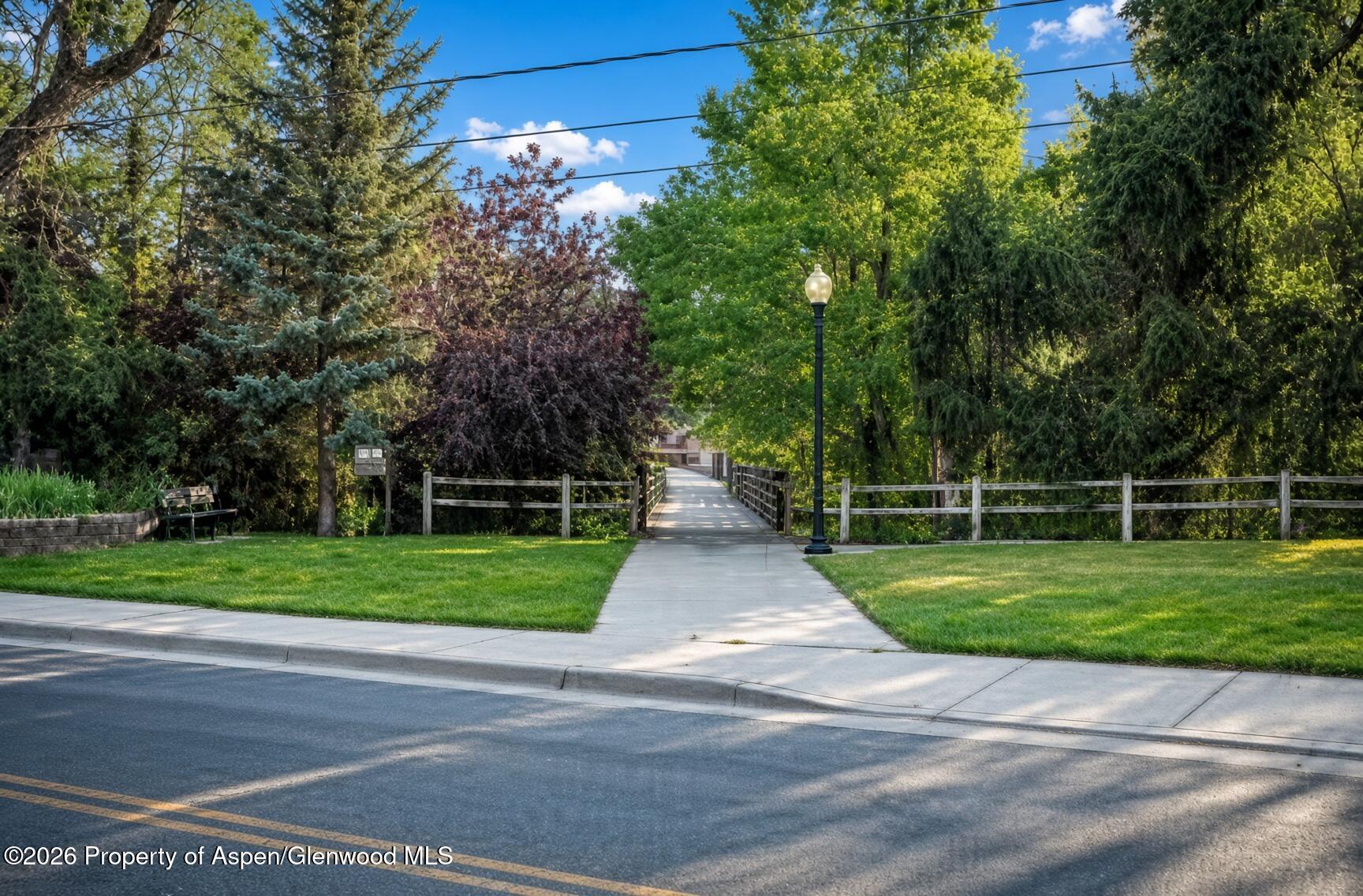 Tree-lined pedestrian path crossing a small bridge through a landscaped neighborhood green space.