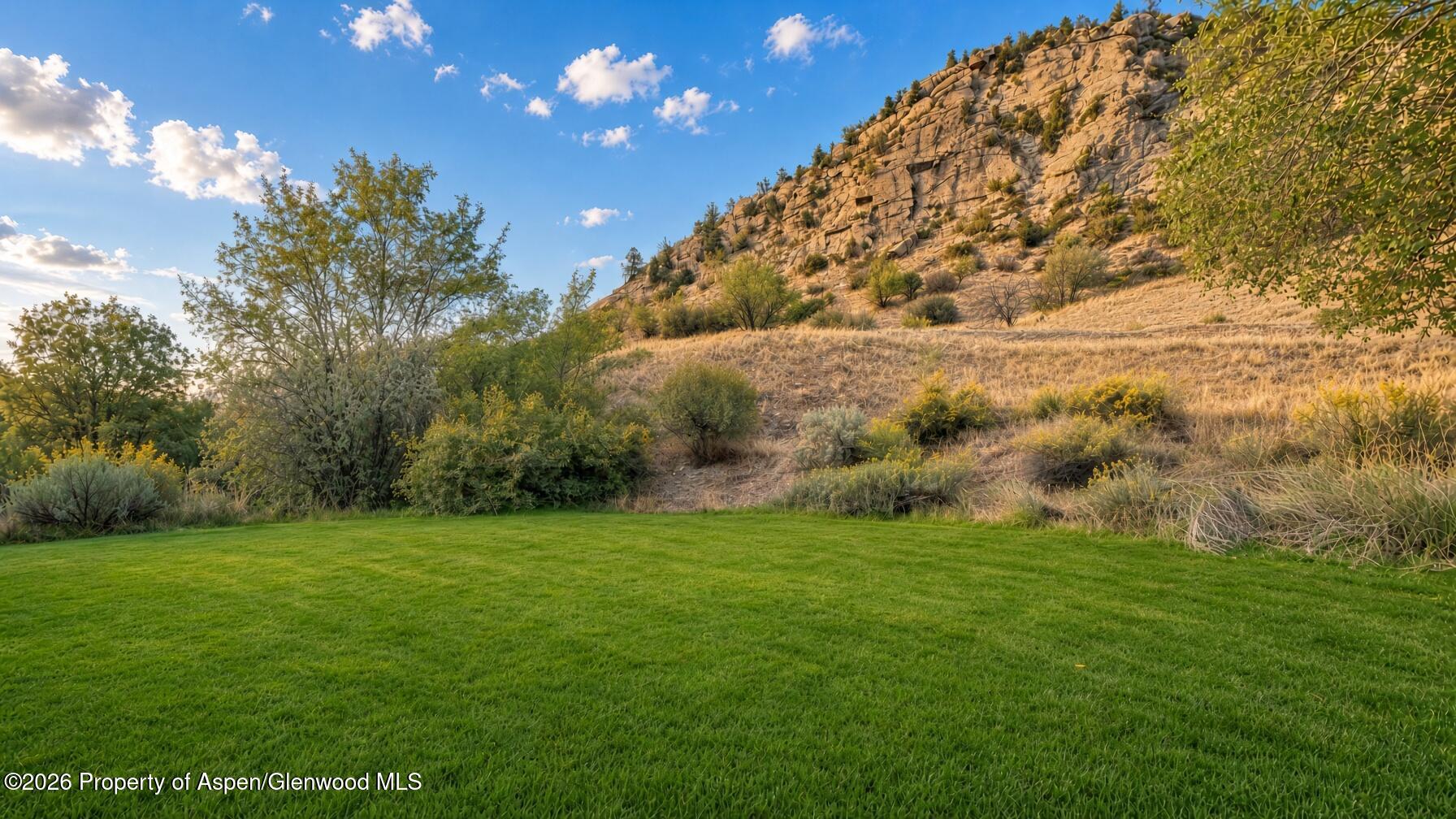 Green lawn beneath a shrub-covered rocky hillside and blue sky.