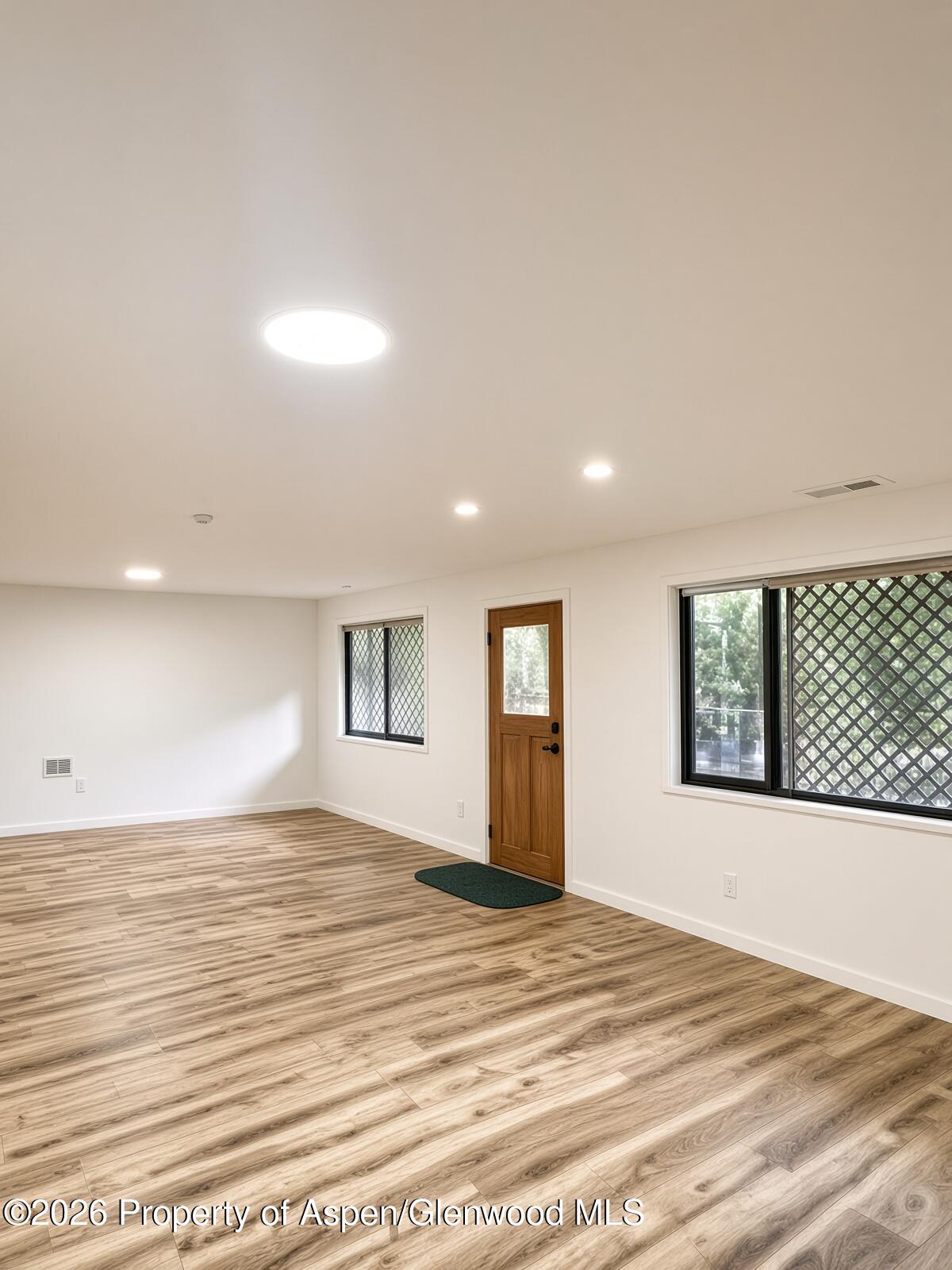Empty room with wood-look plank flooring, recessed lights, and a wooden door beside lattice-screened windows.