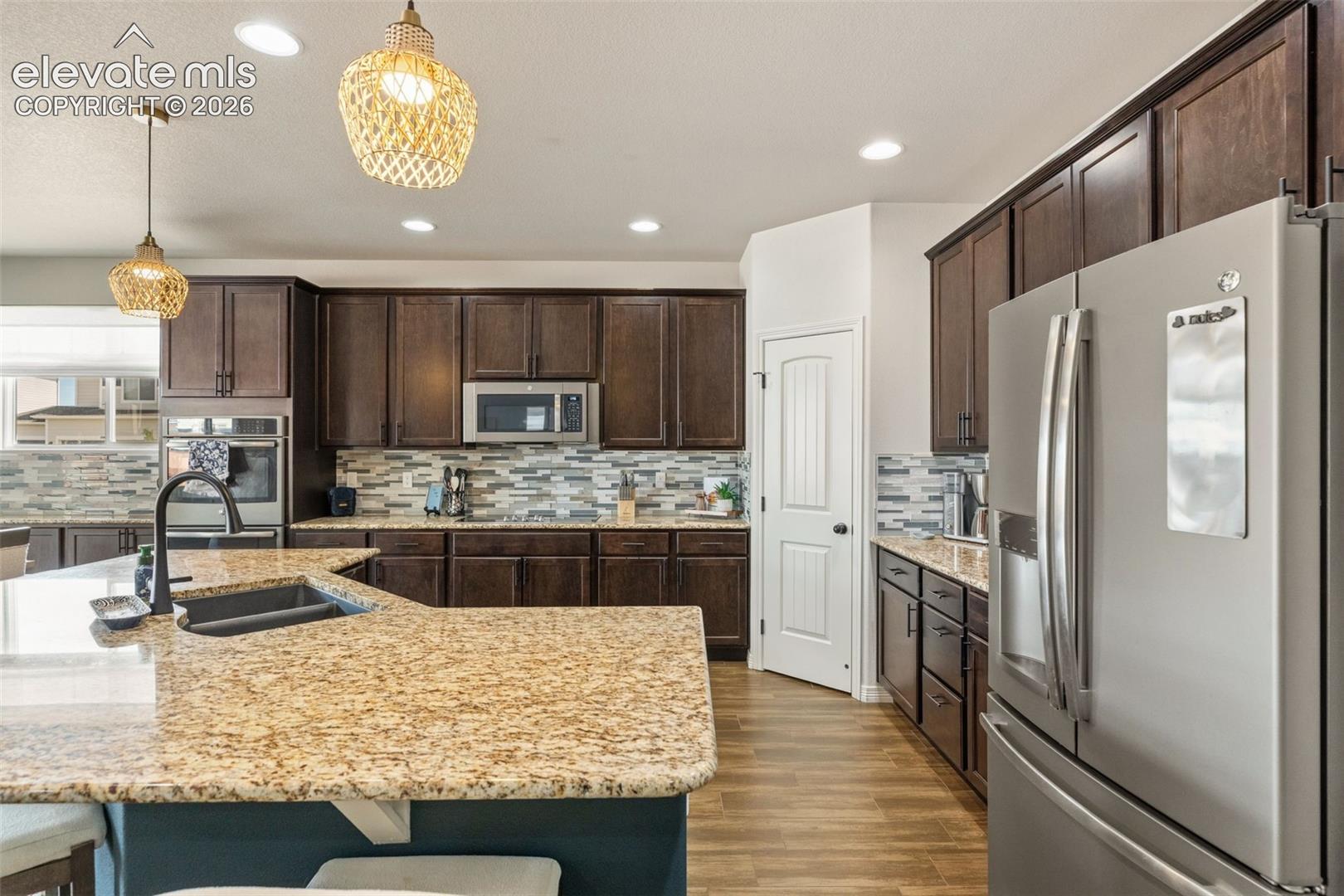 Kitchen with dark wood cabinets, granite island, mosaic backsplash, and stainless steel appliances.