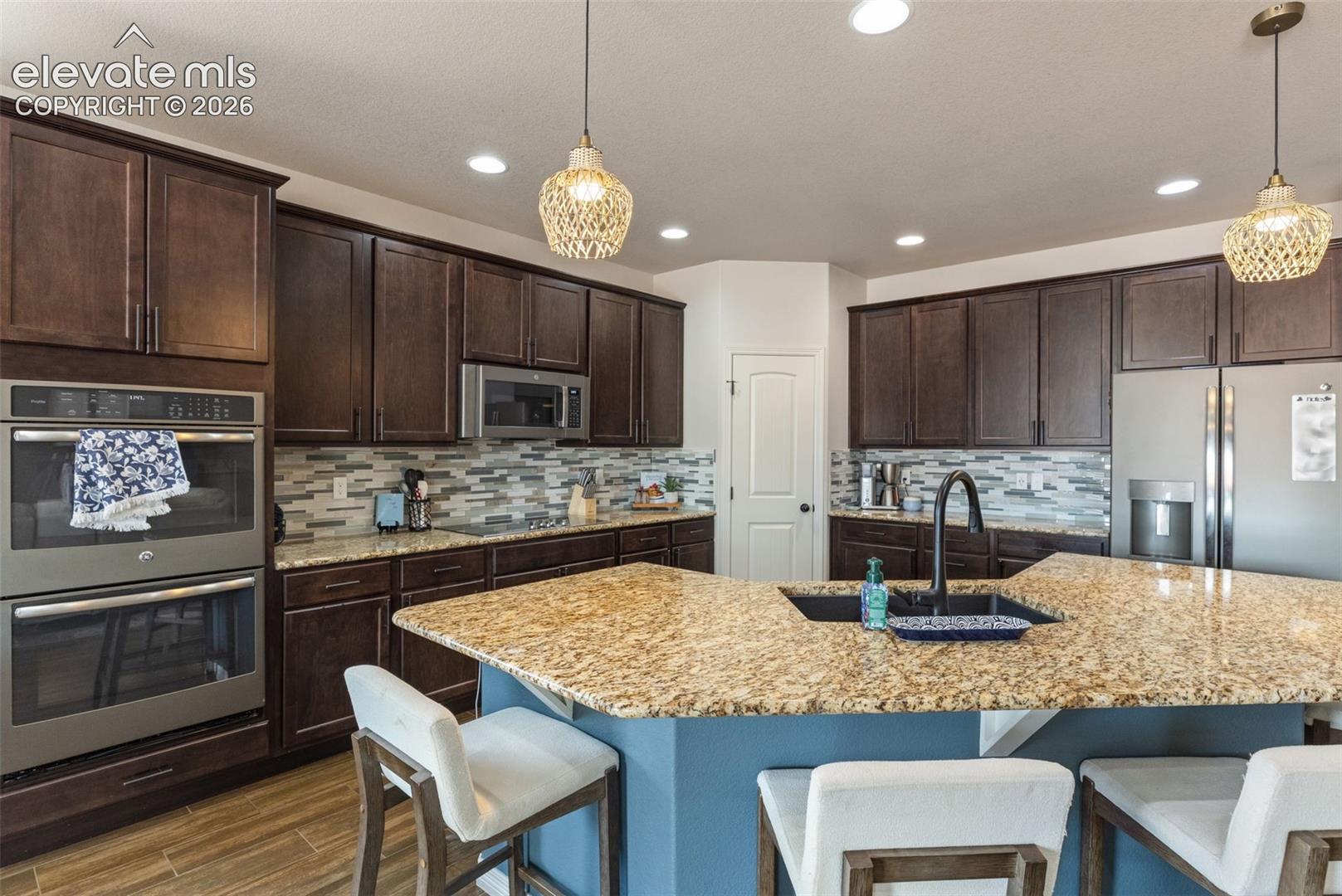 Kitchen with dark wood cabinets, granite island, stainless steel appliances, and mosaic tile backsplash.