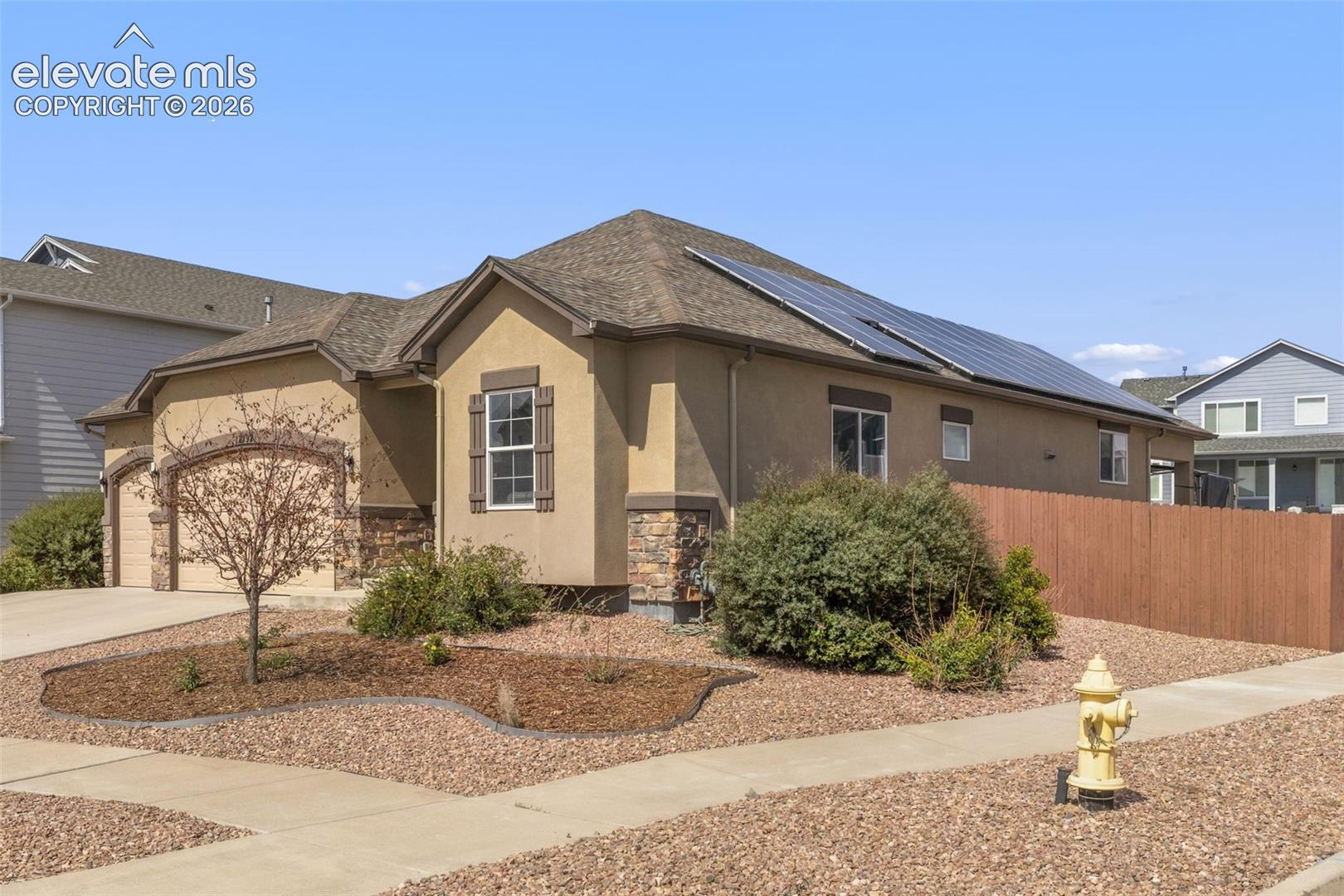 Tan stucco home with rooftop solar panels, stone accents, and a gravel front yard.