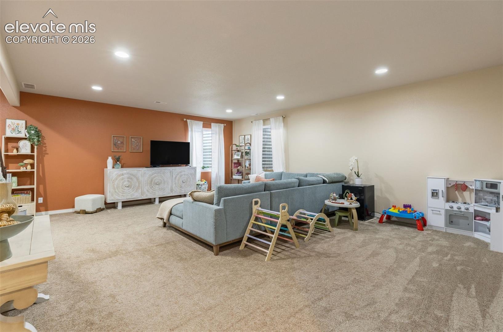 Furnished living room with a gray sectional, rust-colored accent wall, and children’s play area.