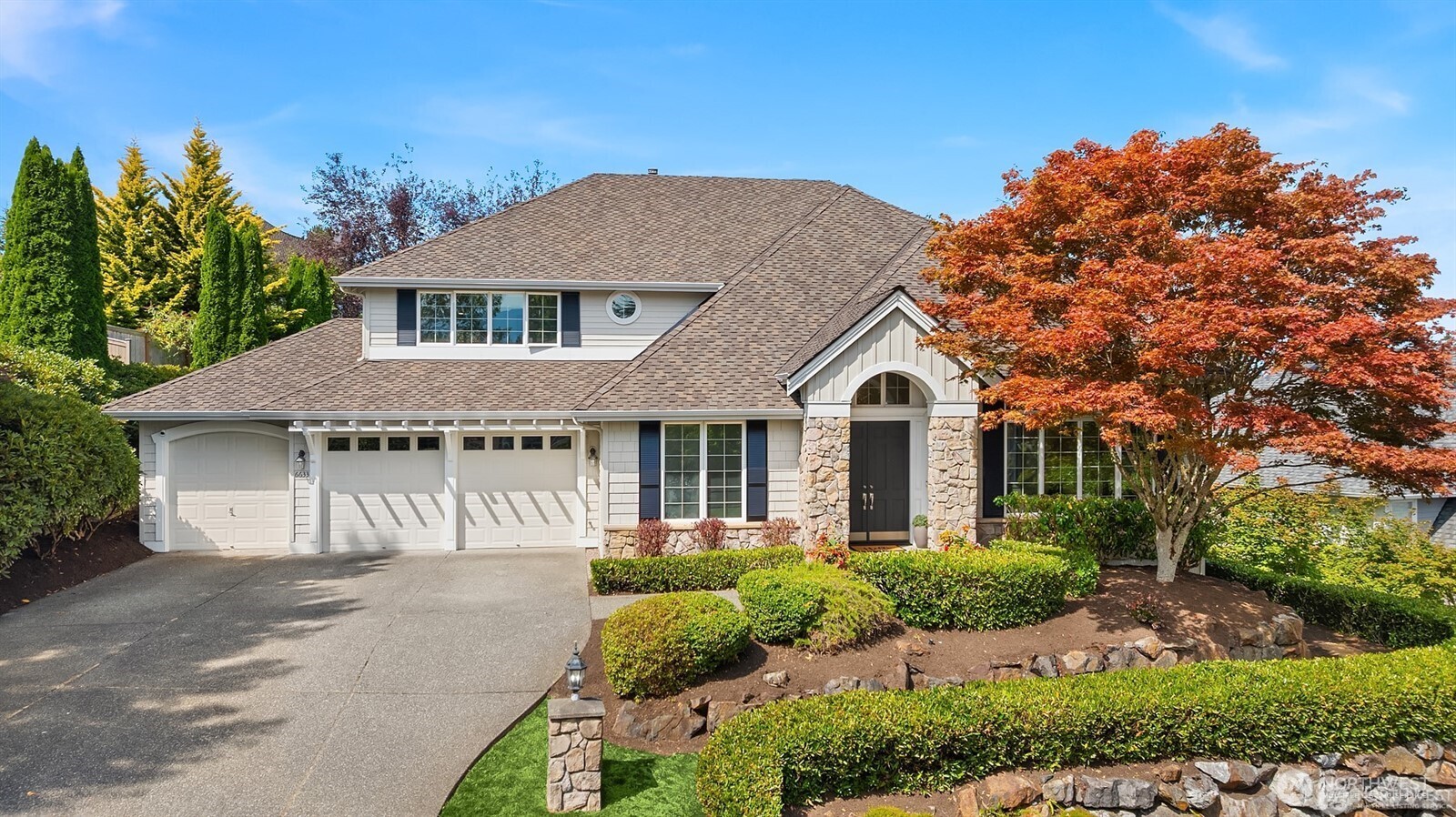 Two-story home with a three-car garage, stone entry, and red-orange maple in the landscaped front yard.