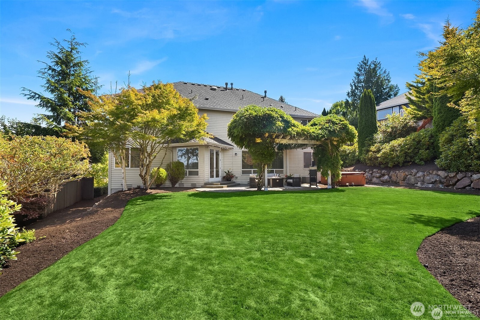 Back of a two-story home with a broad lawn, vine-covered pergola, and hot tub.