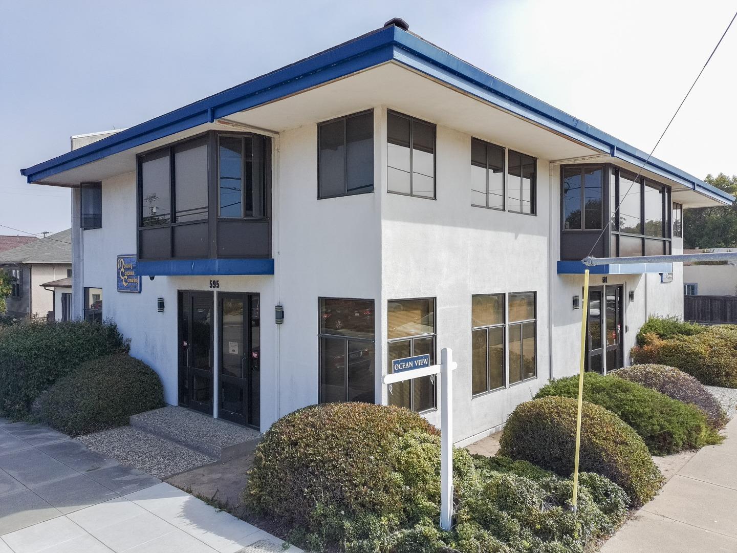 Two-story office building with blue roof trim, broad windows, and landscaped shrubs.