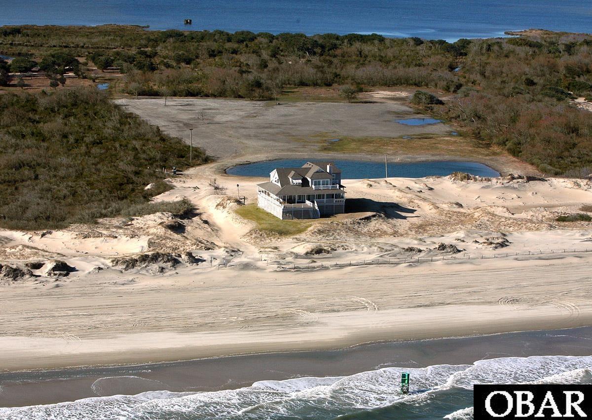 Aerial view of a coastal house on a sandy beach, with dunes, a pond, and ocean surf.