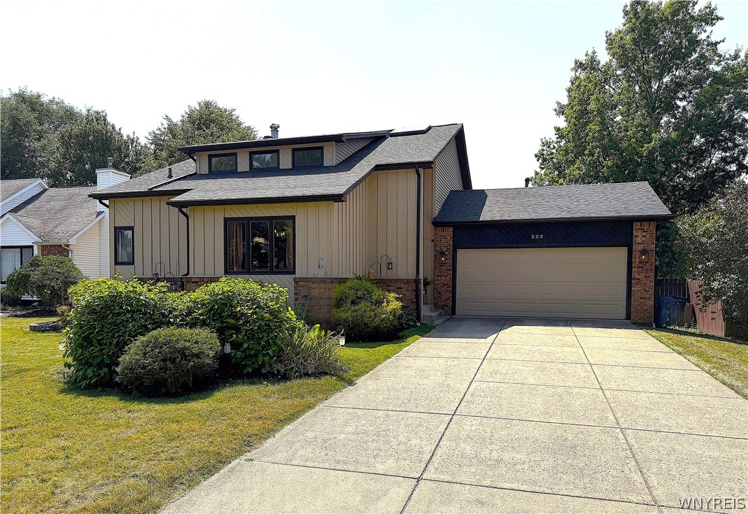 Tan two-story house with brick accents, two-car garage, wide driveway, and landscaped shrubs.