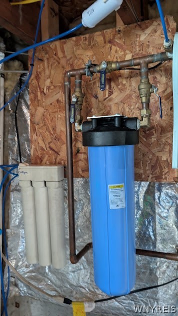 Water filtration system with a tall blue canister, white filter cartridges, and exposed plumbing.