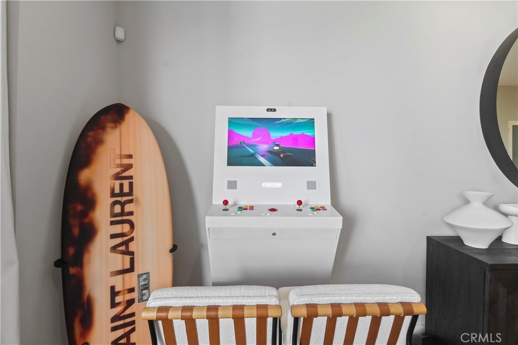 Interior corner with a white arcade cabinet, upright surfboard, and two wooden chairs.