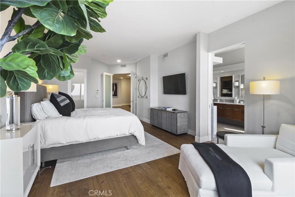 Furnished bedroom with a white bed, hardwood floors, wall-mounted TV, and doorway to a bathroom.