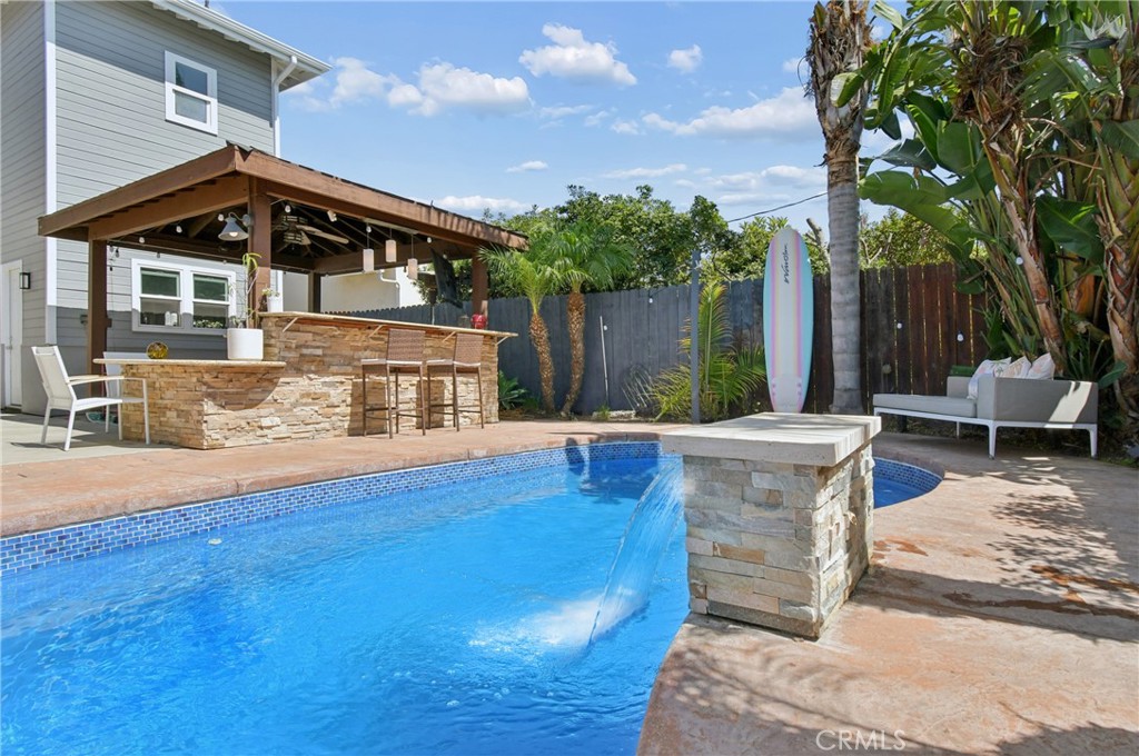 Backyard pool beside a covered outdoor kitchen, stone counter, and tropical landscaping.