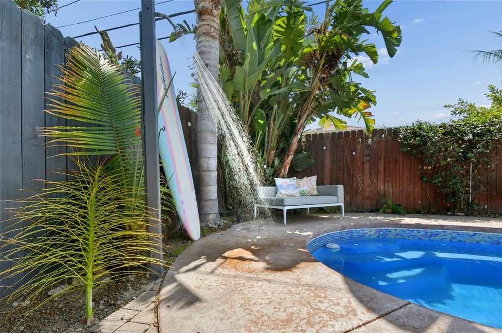 Backyard pool with a white bench, tropical plants, and a surfboard beside the patio.