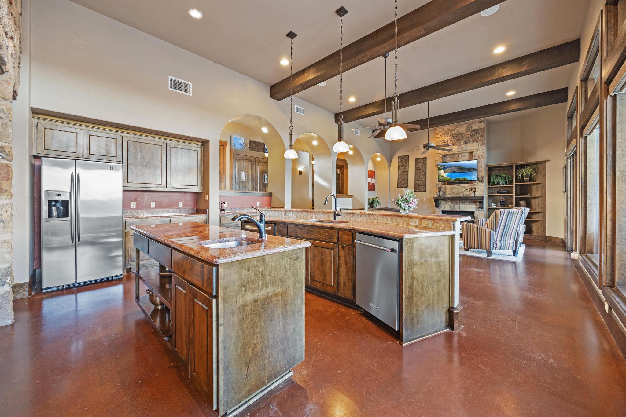 Spacious kitchen with two stone-topped islands, exposed wood beams, and arched openings to a living area.