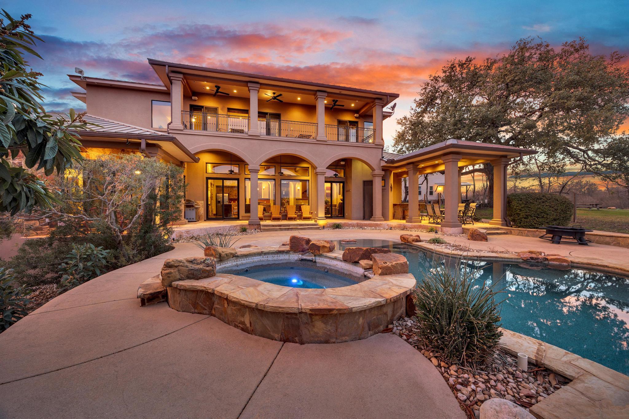 Two-story stucco home with arched patio, balcony, swimming pool, and raised circular spa at sunset.