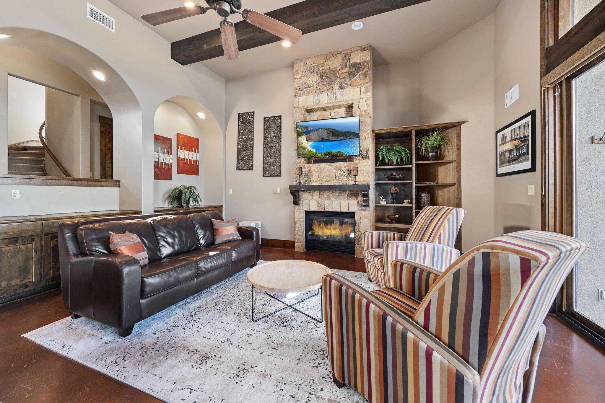 Furnished living room with a stone fireplace, exposed beams, leather sofa, and striped armchairs.