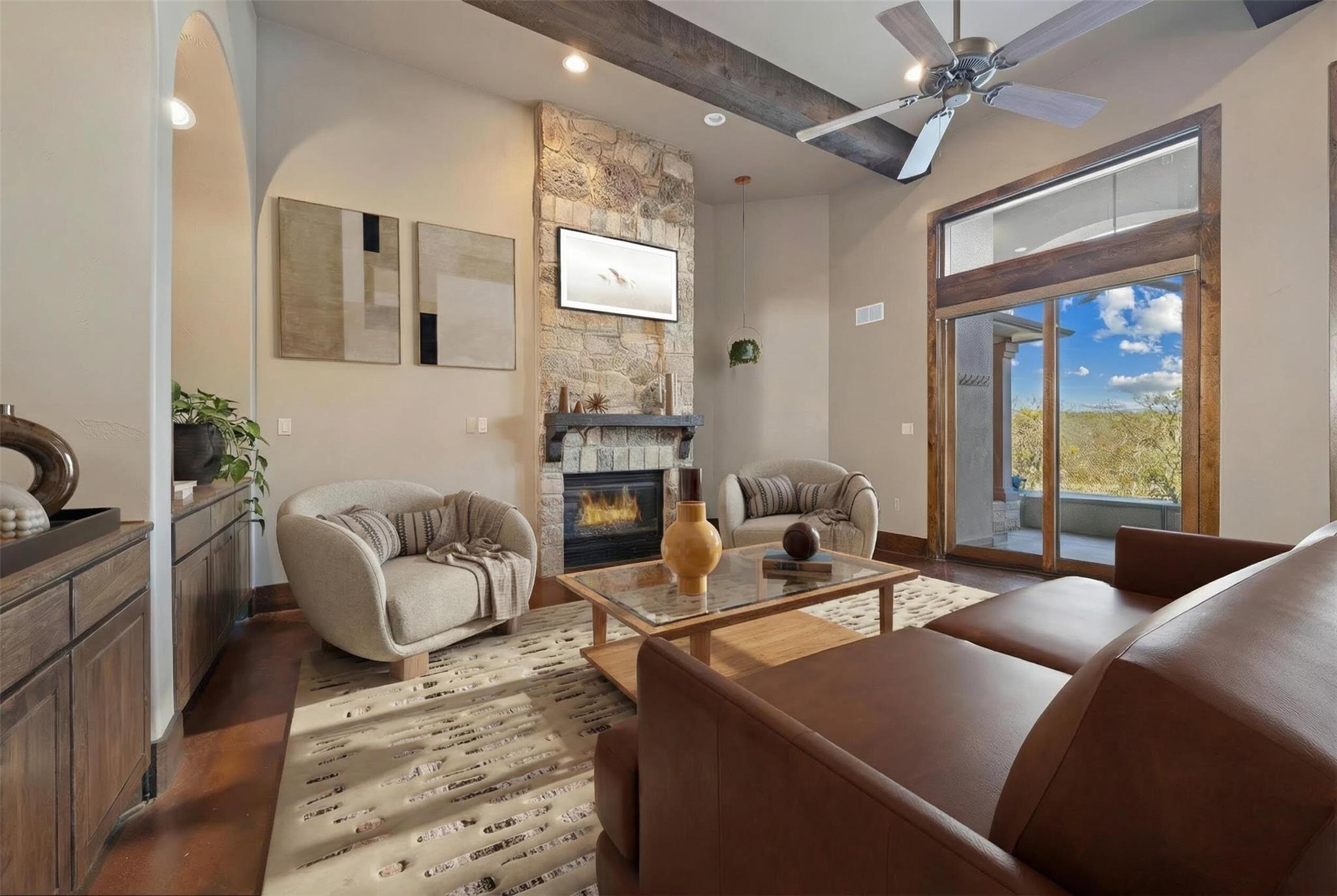 Living room with a stone fireplace, exposed beam, and sliding doors overlooking trees.
