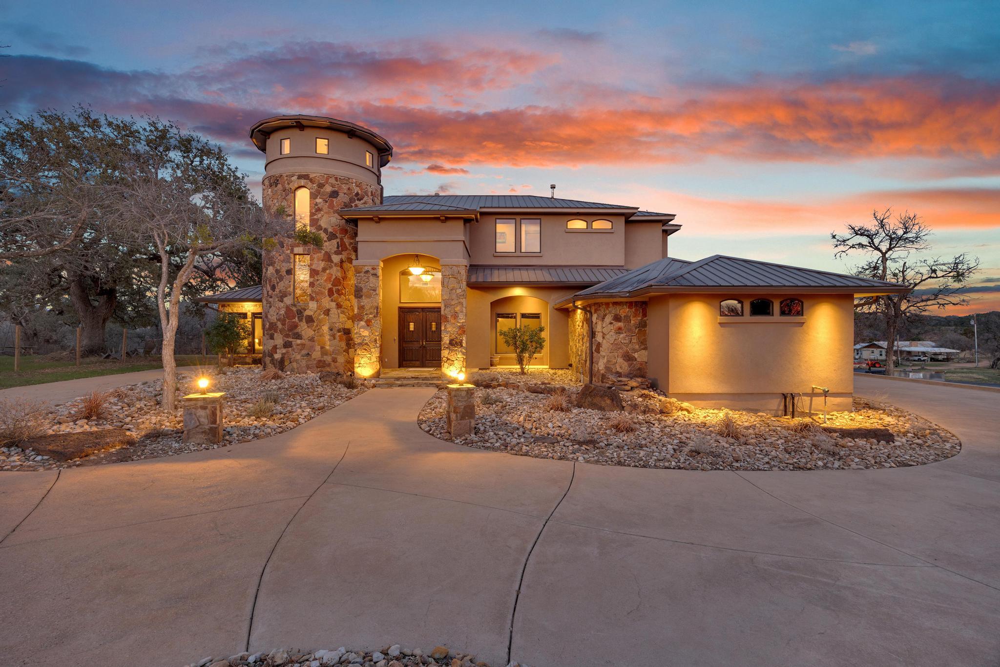 Two-story home with a round stone tower and illuminated landscaping at dusk.