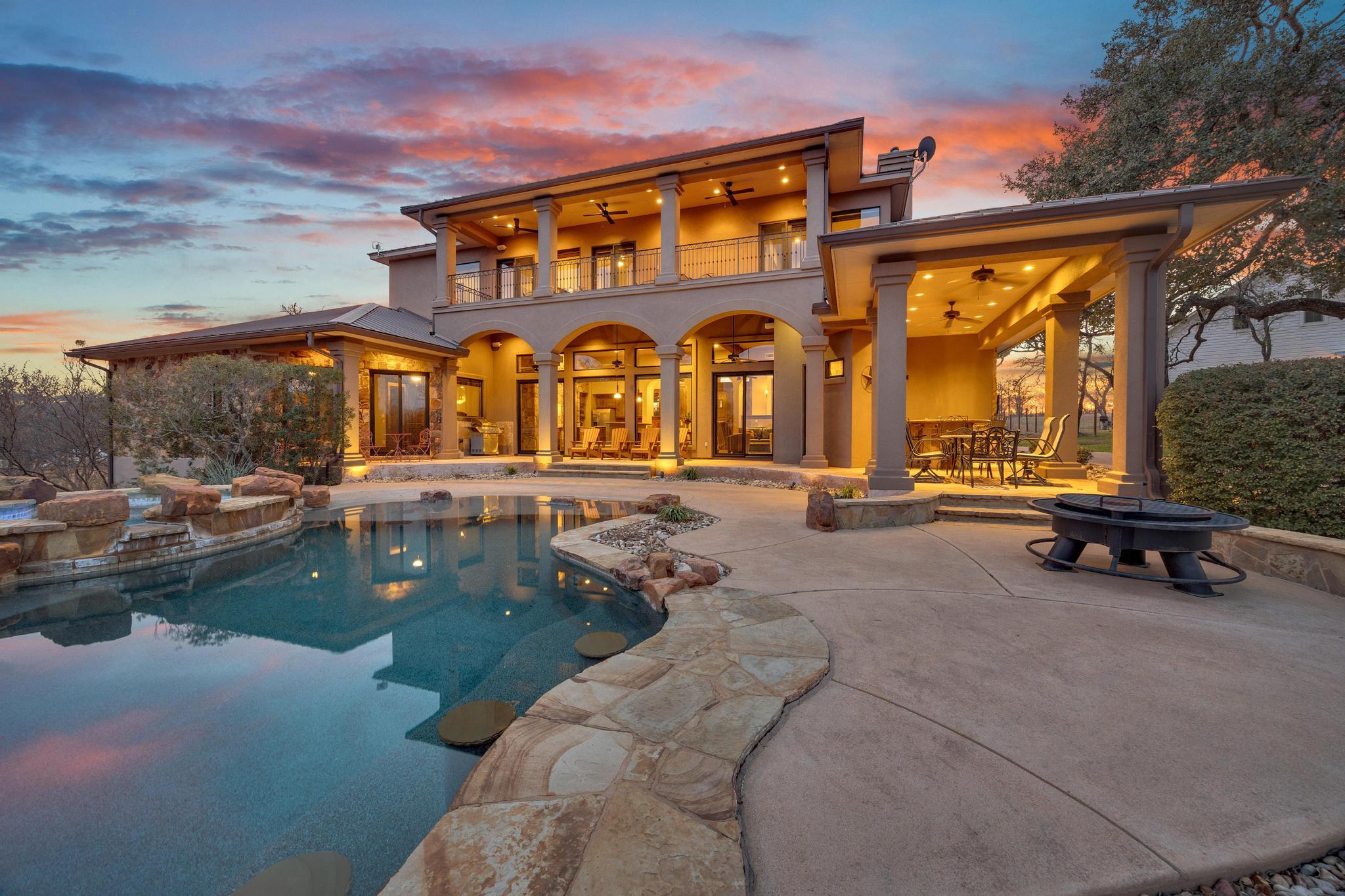 Two-story home with an illuminated arched patio and balcony beside a stone-edged pool at dusk.