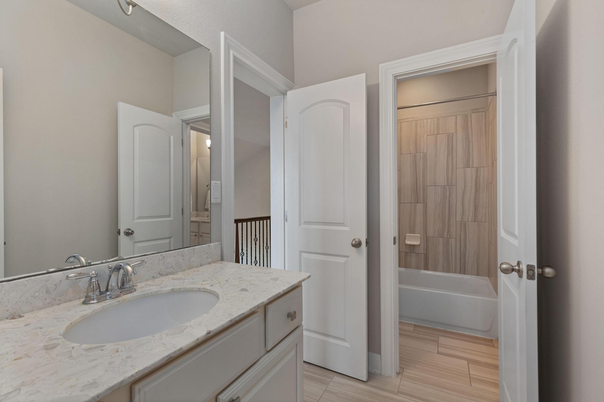 Bathroom vanity with pale stone countertop beside a tub with wood-look tile surround.