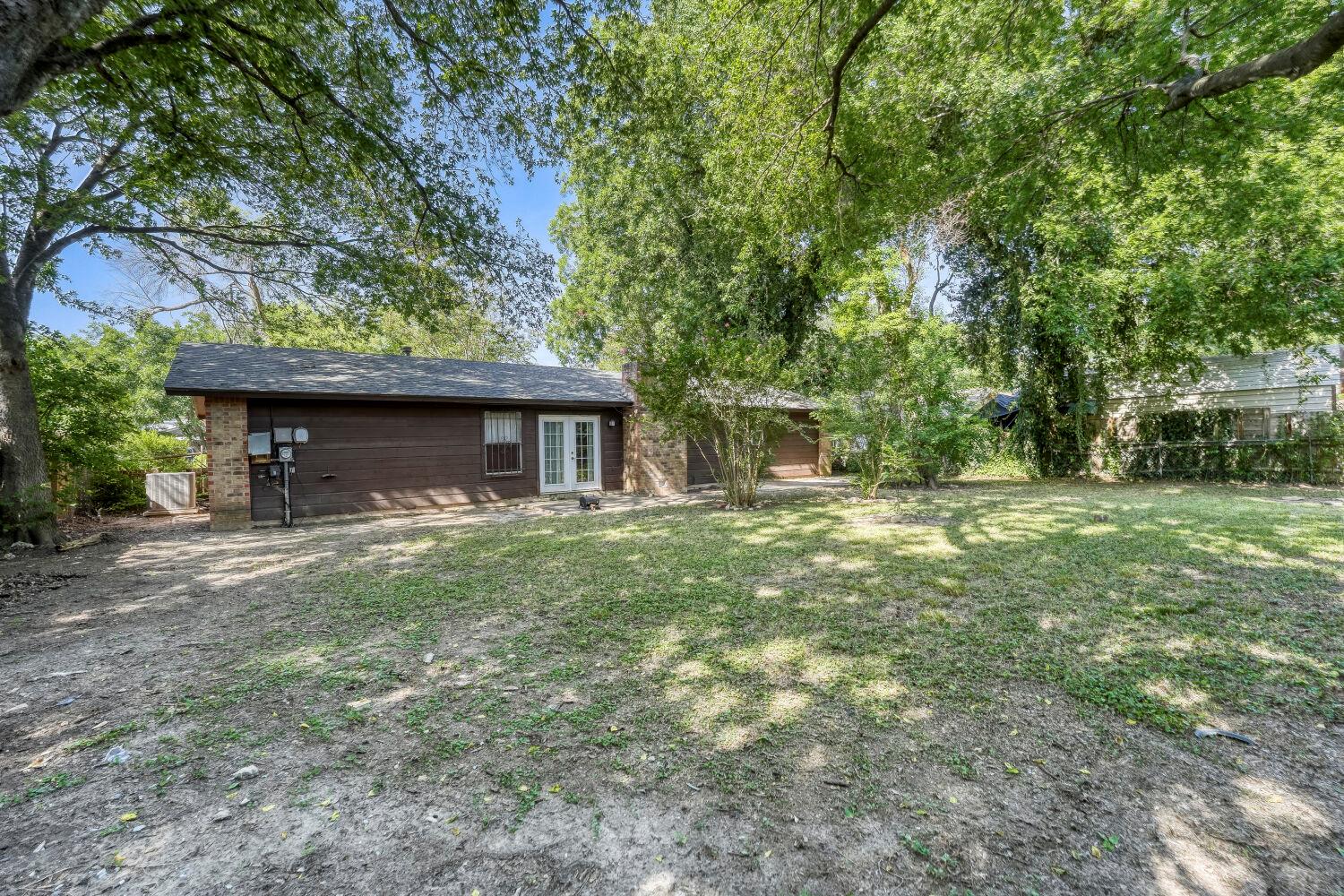Single-story brown house with brick accents and French doors beneath mature trees.