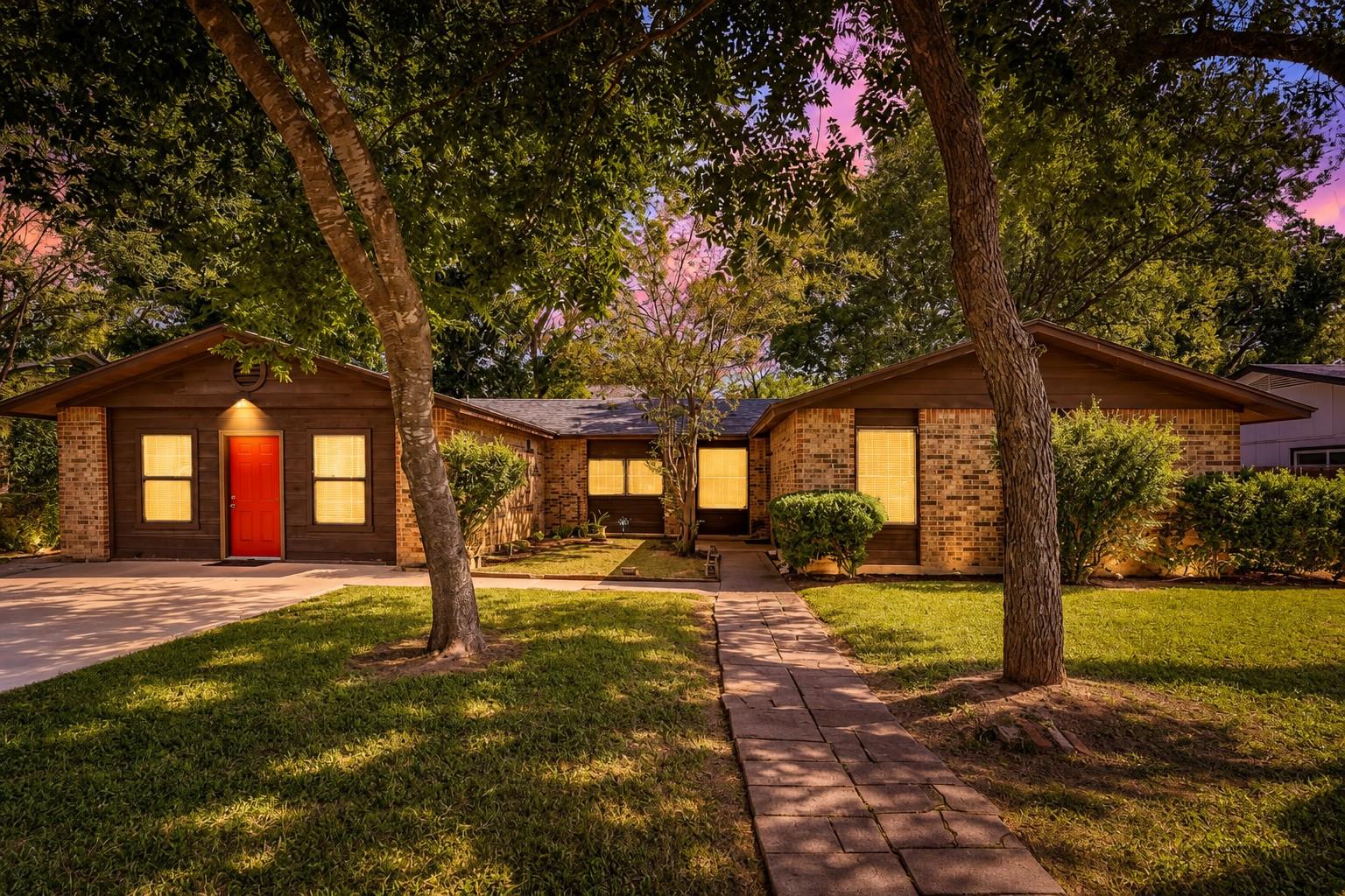 Single-story brick home with a bright red front door, mature trees, and a stone walkway.