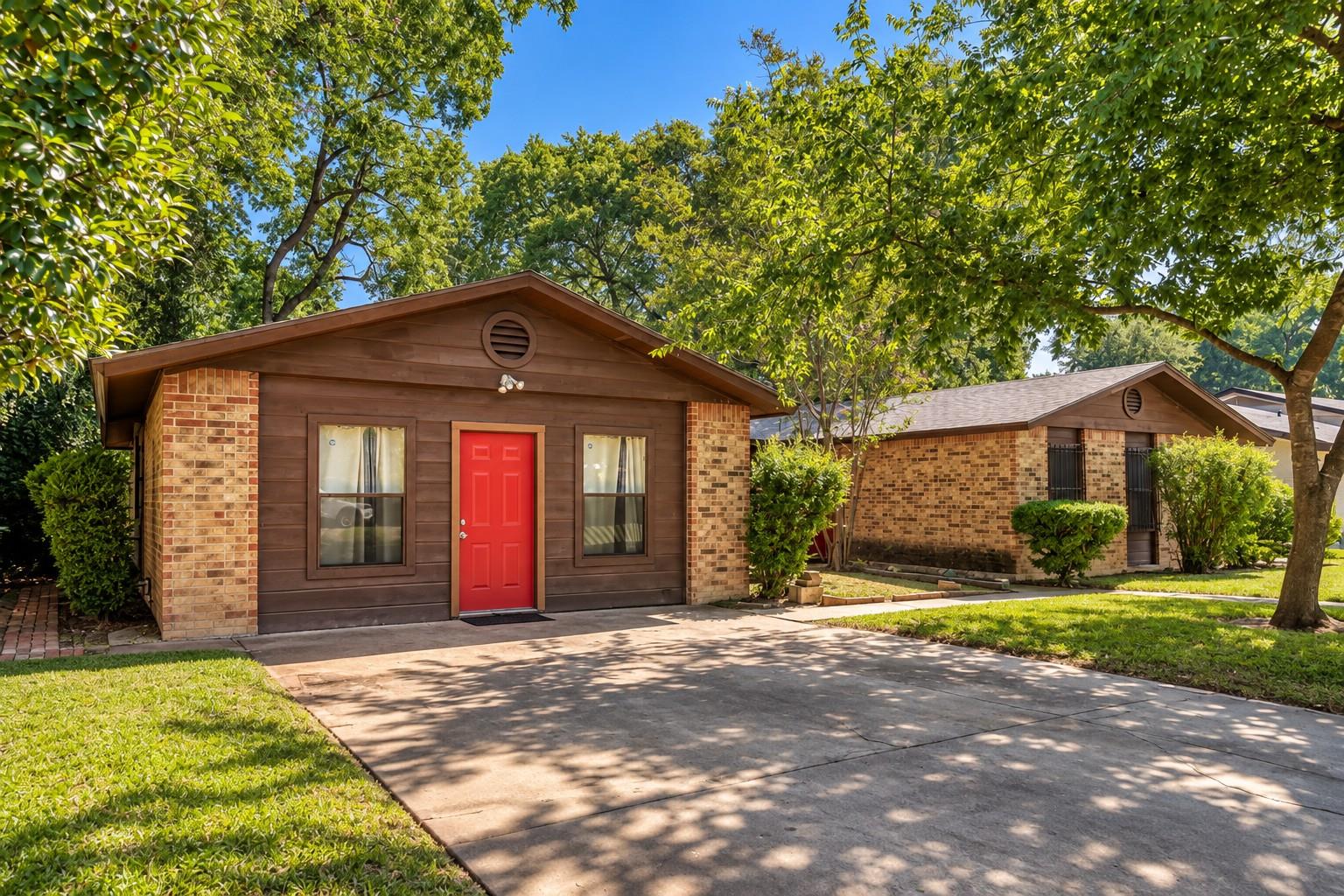 Single-story brown-and-brick home with a bright red front door beneath mature trees.