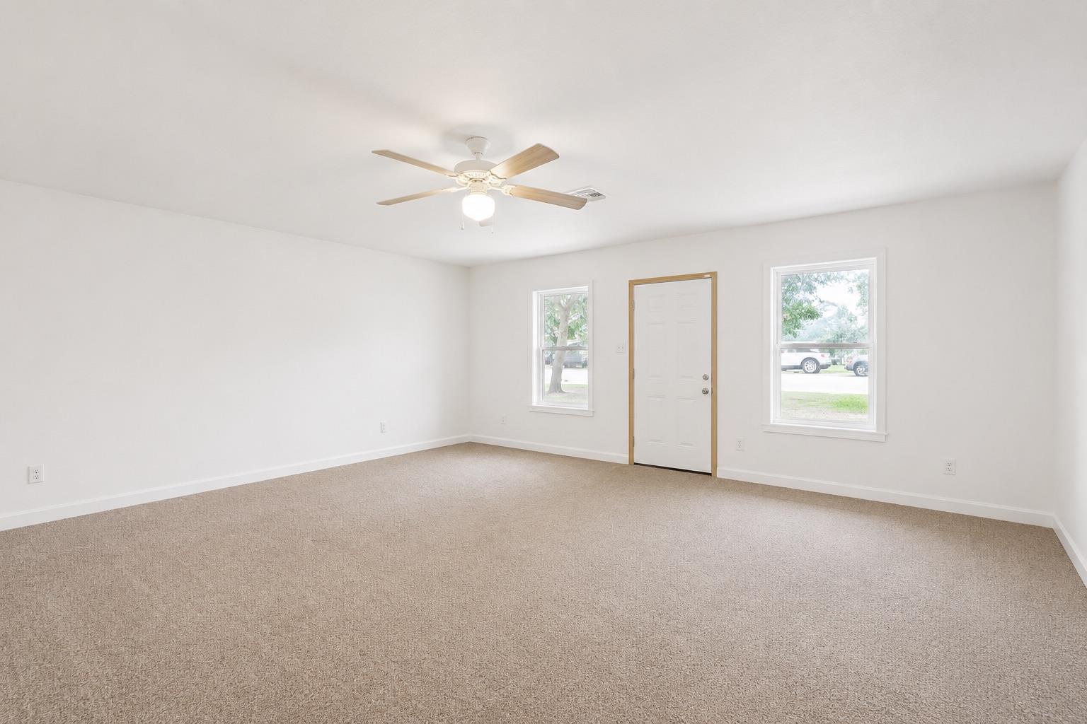 Vacant carpeted room with a ceiling fan and an exterior door between two windows.