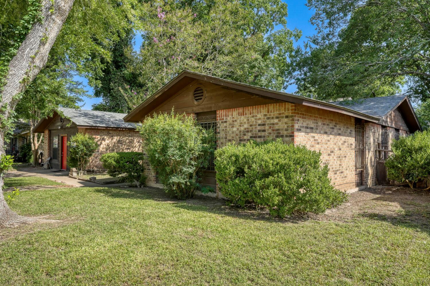 Brick house with intersecting gabled roofs, red front door, mature trees, and broad lawn.