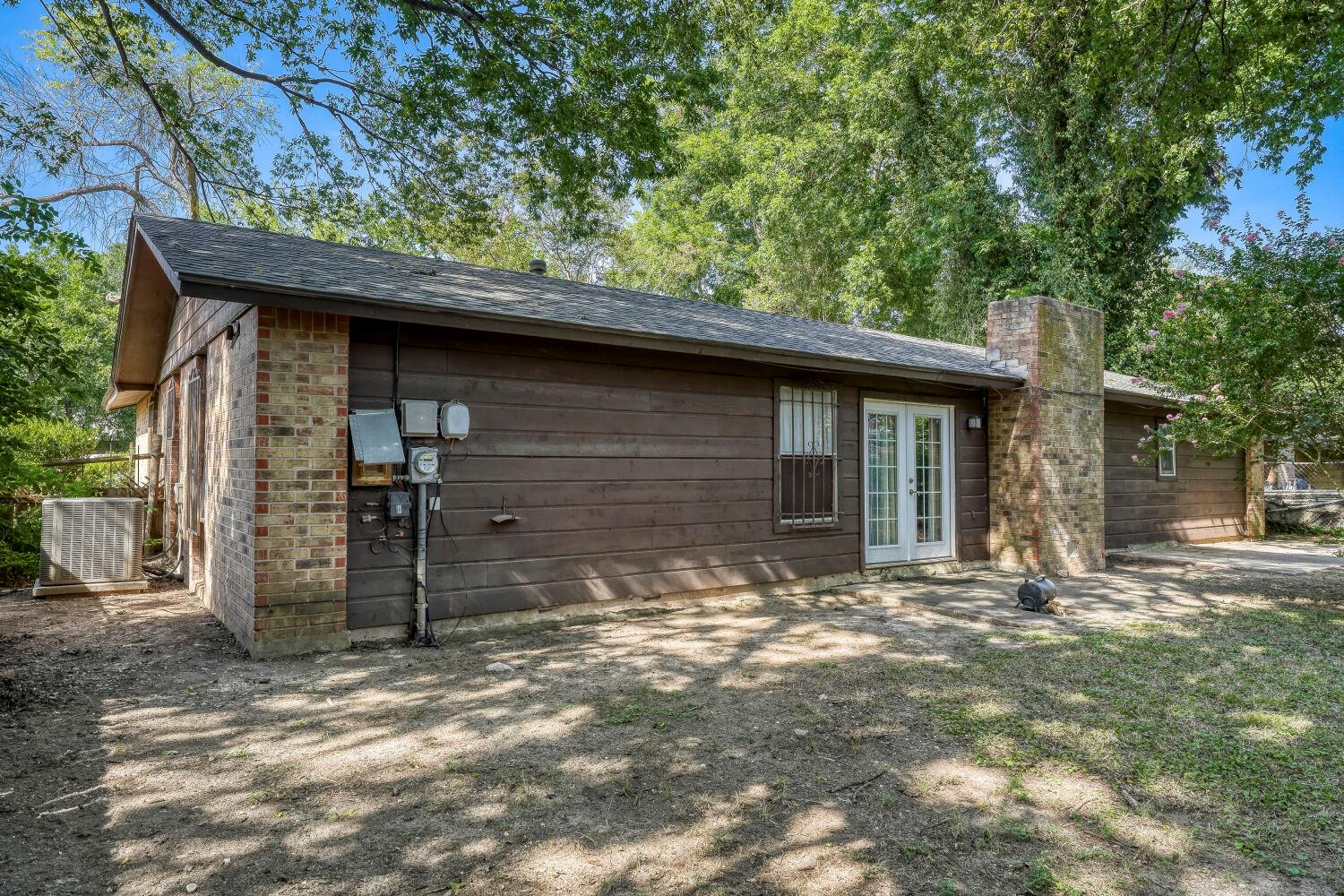 Single-story house with brown siding, brick chimney, and French doors beneath mature trees.