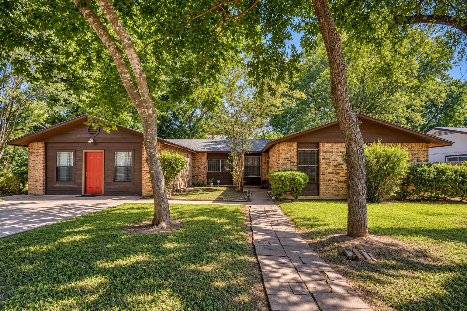 Single-story brick house with a red front door, mature shade trees, and a stone walkway.