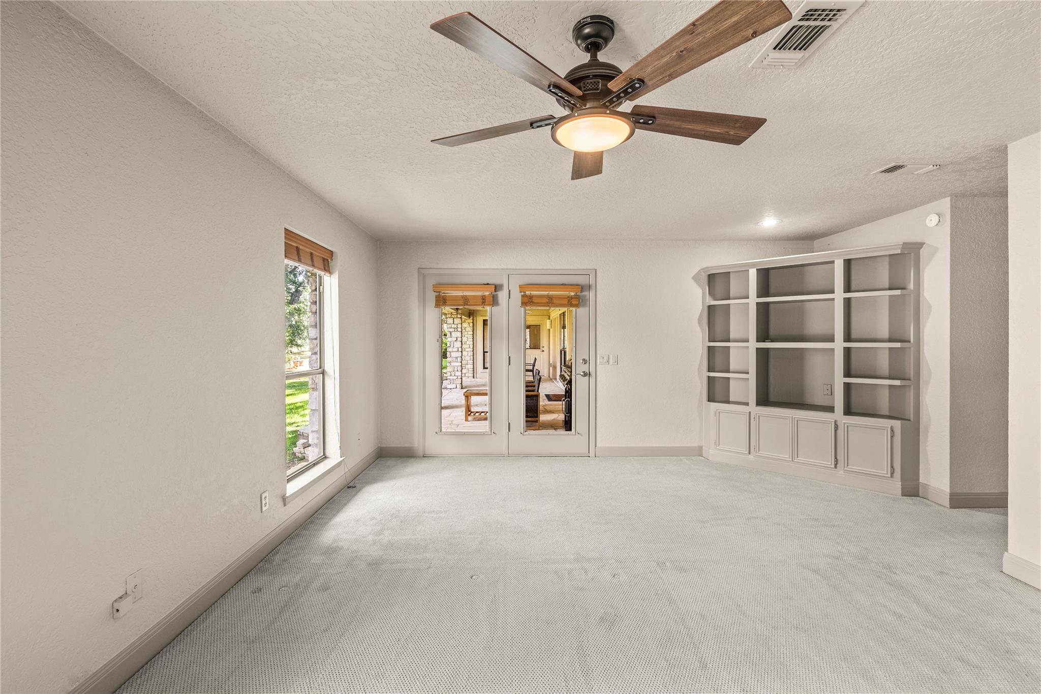 Empty carpeted room with built-in shelves, a ceiling fan, and glass doors to a patio.