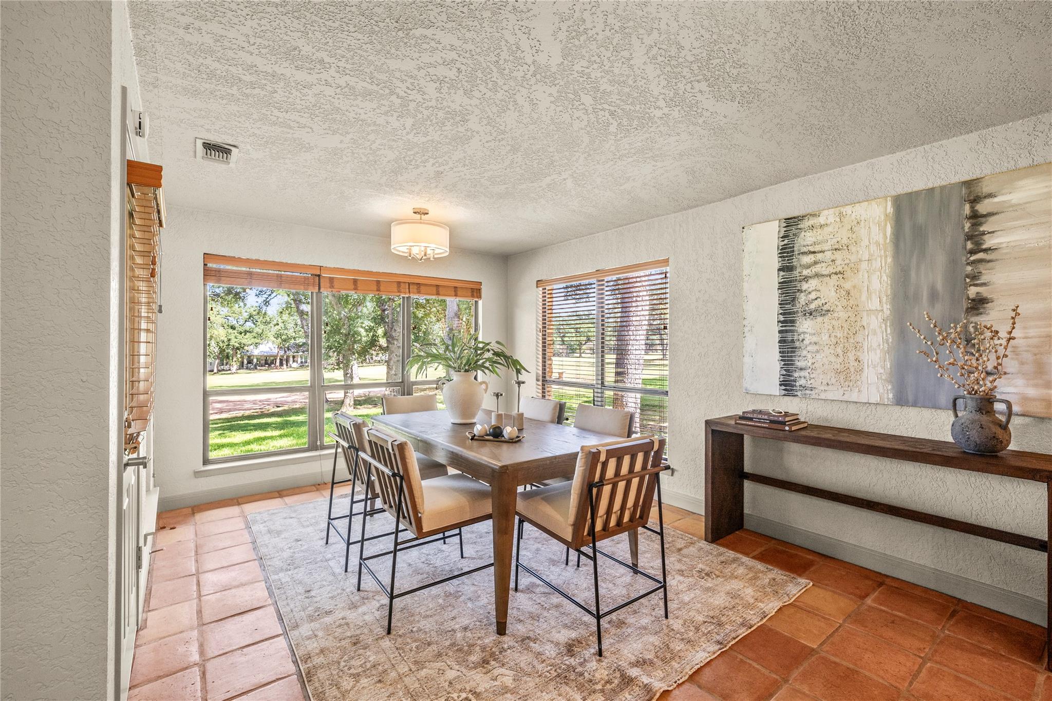 Dining room with a wood table and chairs, terracotta tile floor, and large windows overlooking a green lawn.