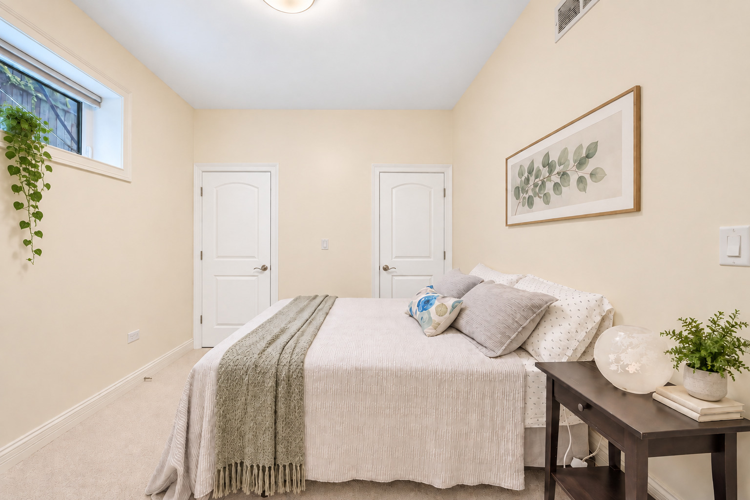 Staged bedroom with a high window, trailing plant, and light bed with a gray knitted throw.