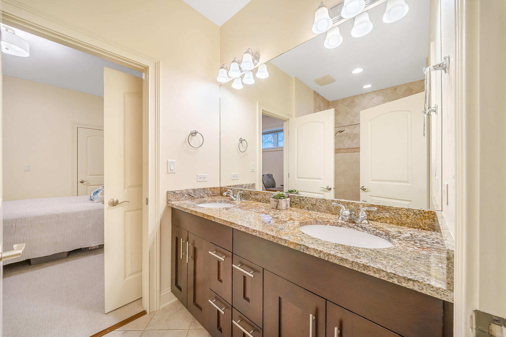 Bathroom with a granite double-sink vanity and dark wood cabinetry, adjoining a bedroom.